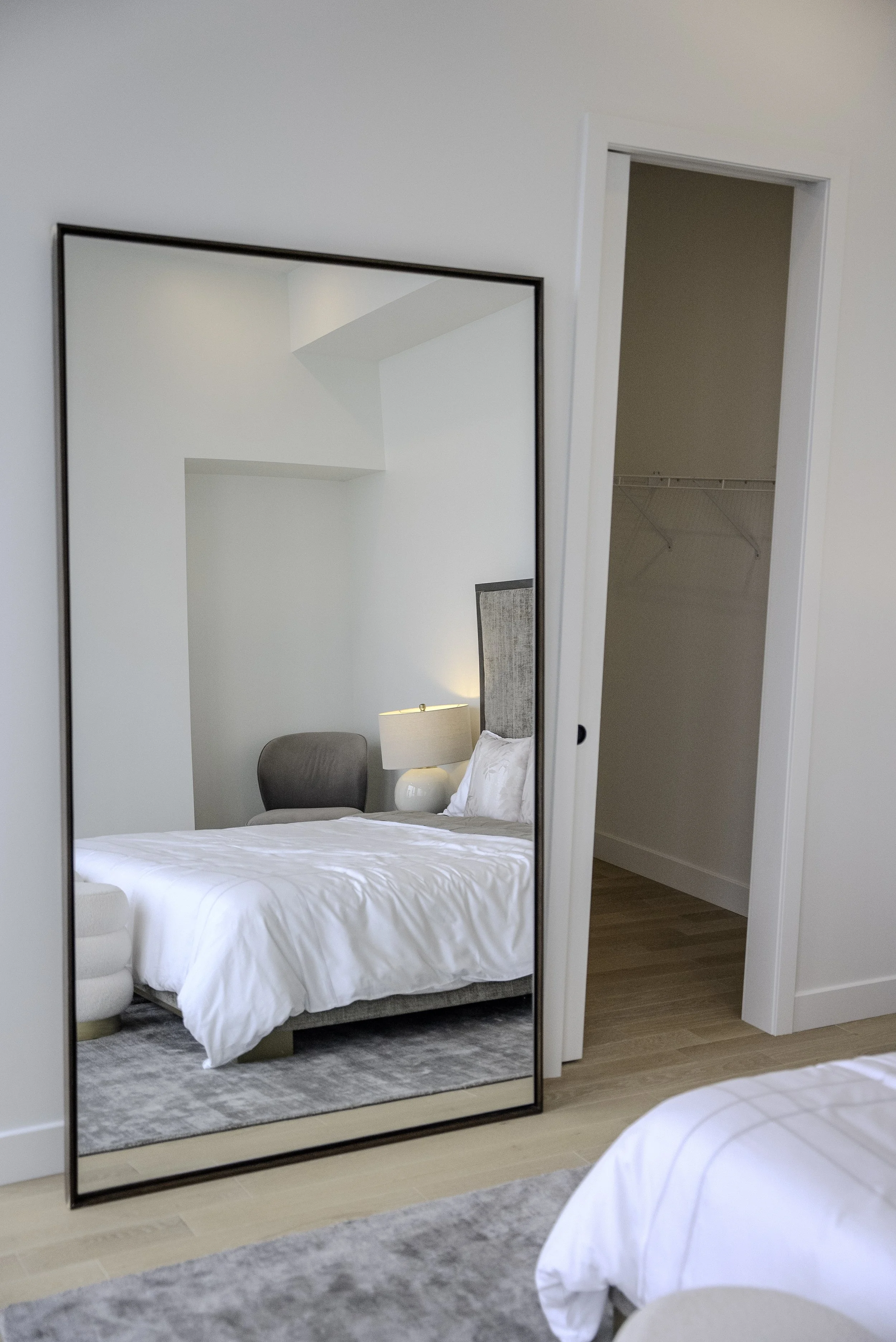 Un miroir refletant un lit avec des draps blancs, une lampe de chevet et une chaise. On voit aussi une armoire ouverte vide. La pièce a un sol en bois clair.