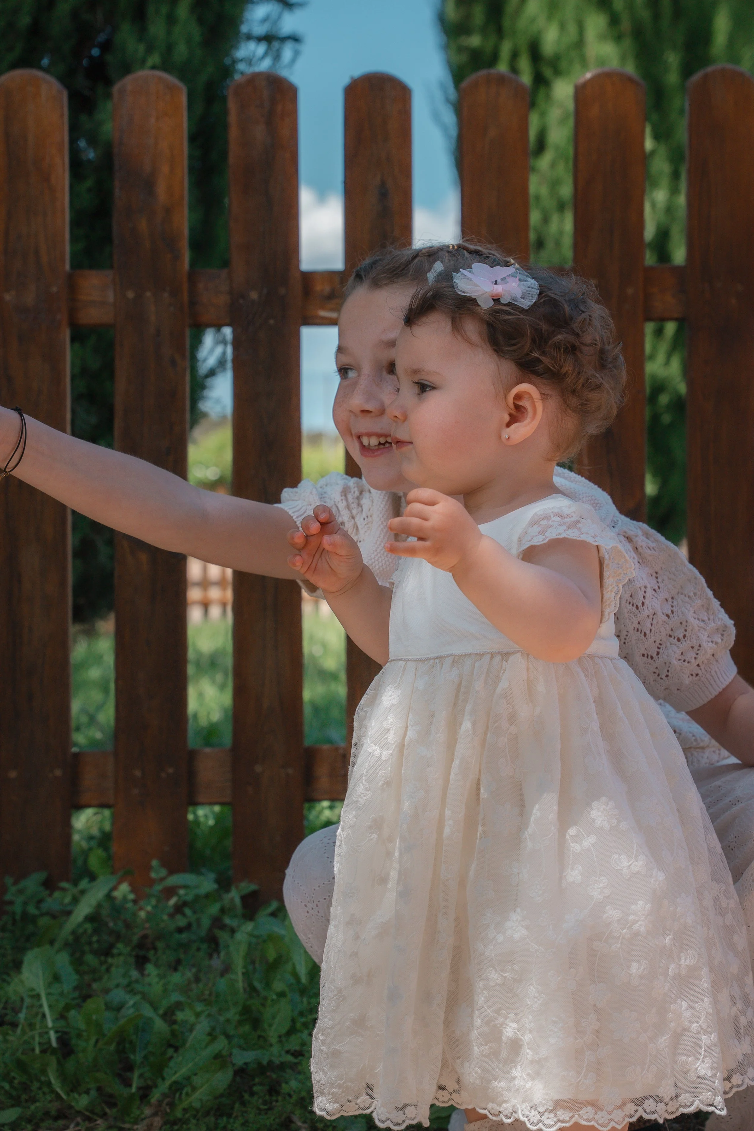 Deux jeunes filles habillées en blanc, l'une plus grande que l'autre, prennent une photo ensemble devant une clôture en bois, avec des arbres verts en arrière-plan, un jour ensoleillé.