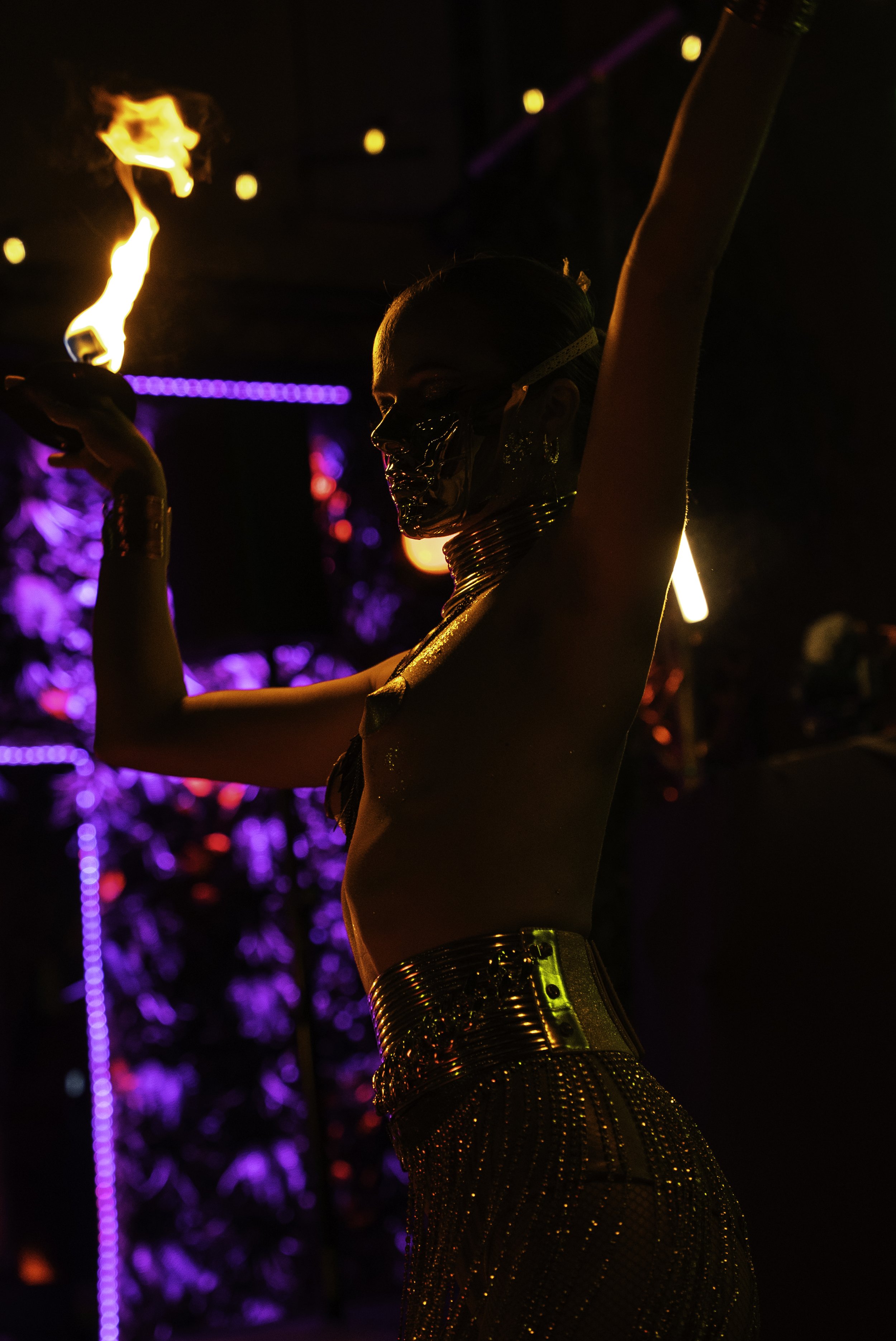 Une femme en costume de spectacle, faisant un numéro avec un feu, dans un décor éclairé à la lumière vive et colorée, lors d'une performance artistique ou un spectacle de nuit.
