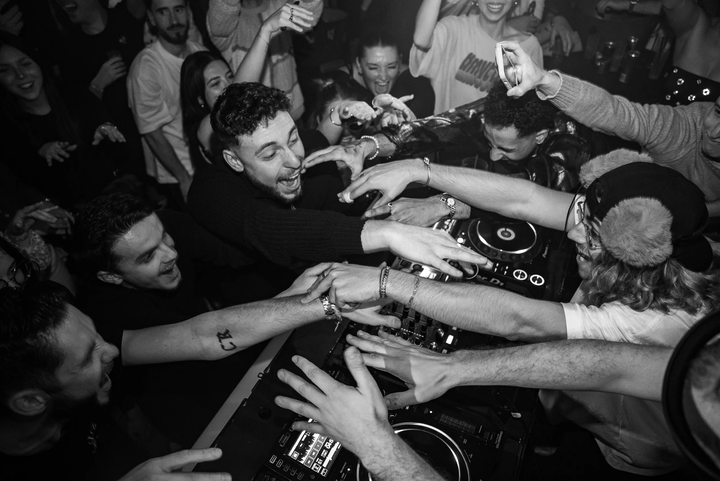 Un DJ en train de mixer de la musique lors d'une fête, entouré de plusieurs personnes qui dansent et profitent de la musique, dans une ambiance animée et joyeuse.