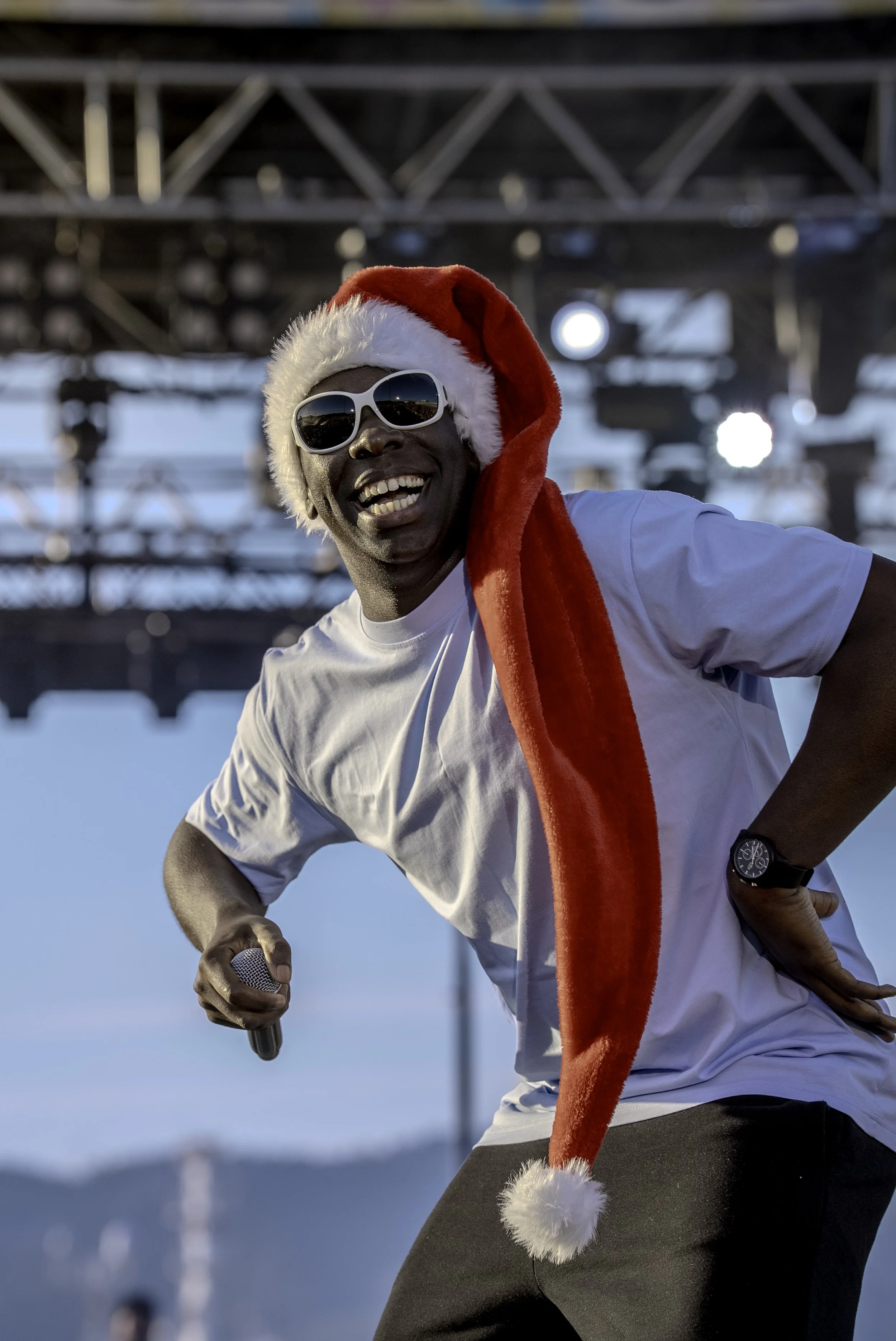 Homme souriant portant un bonnet de Noël, des lunettes de soleil blanches, un tee-shirt blanc, un bracelet noir, tenant un microphone sur scène, sous un ciel bleu.