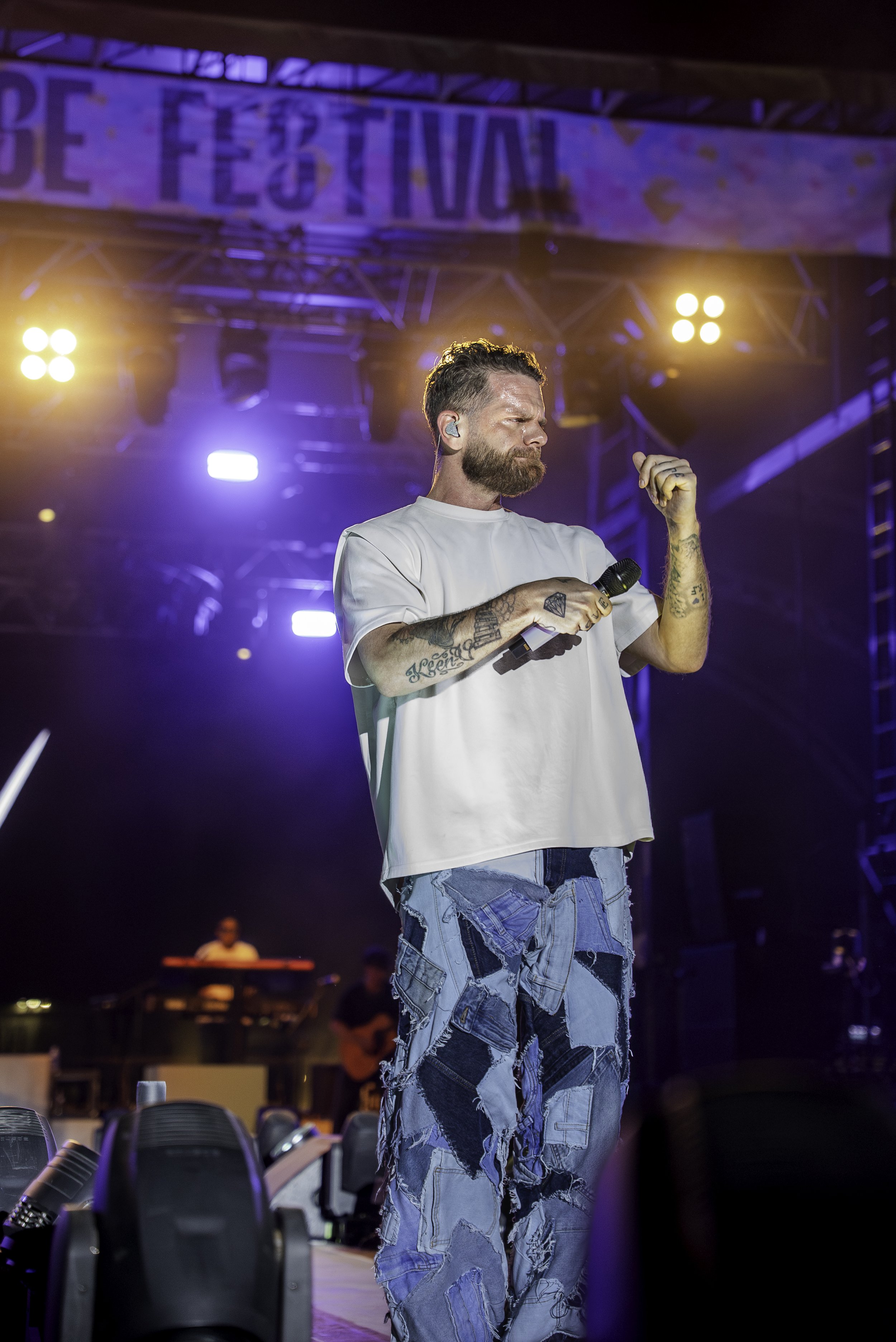 Un homme avec une barbe et des tatouages, portant un t-shirt blanc et un pantalon en jean déchiré, tient un microphone sur scène lors d'un festival, avec des lumières colorées en arrière-plan.