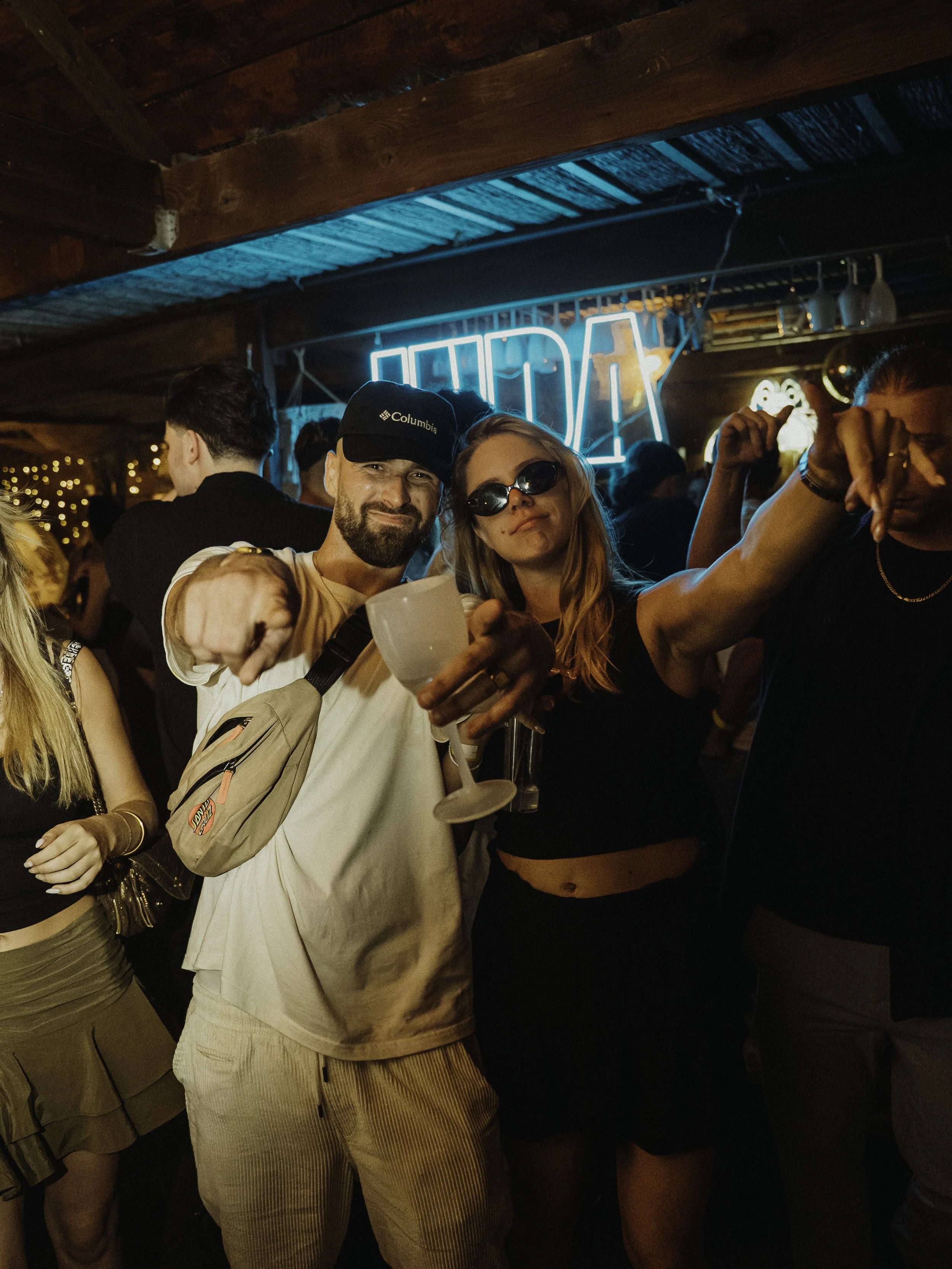 Groupe de jeunes adultes dans une ambiance de fête ou de bar, avec un éclairage néon bleu et des décorations chaleureuses, souriant et levant des verres.
