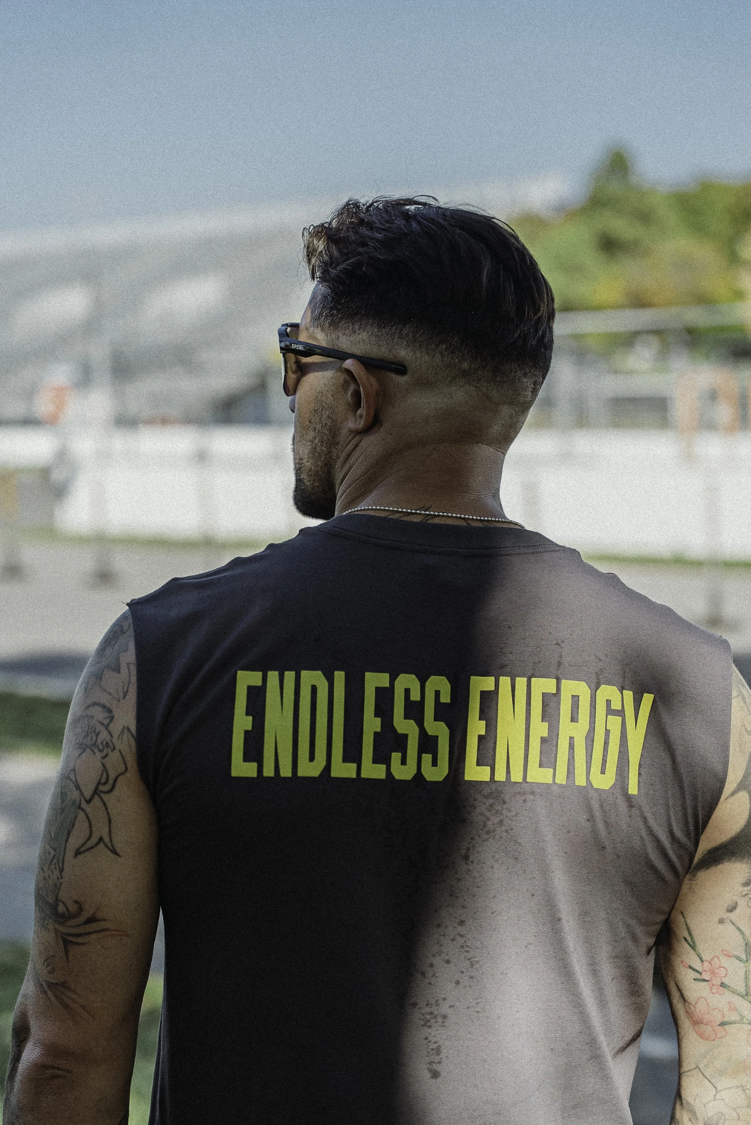 Un homme avec des tatouages, portant un t-shirt noir avec l'inscription "ENDLESS ENERGY" en lettres vertes, vu de dos, dans un environnement extérieur.