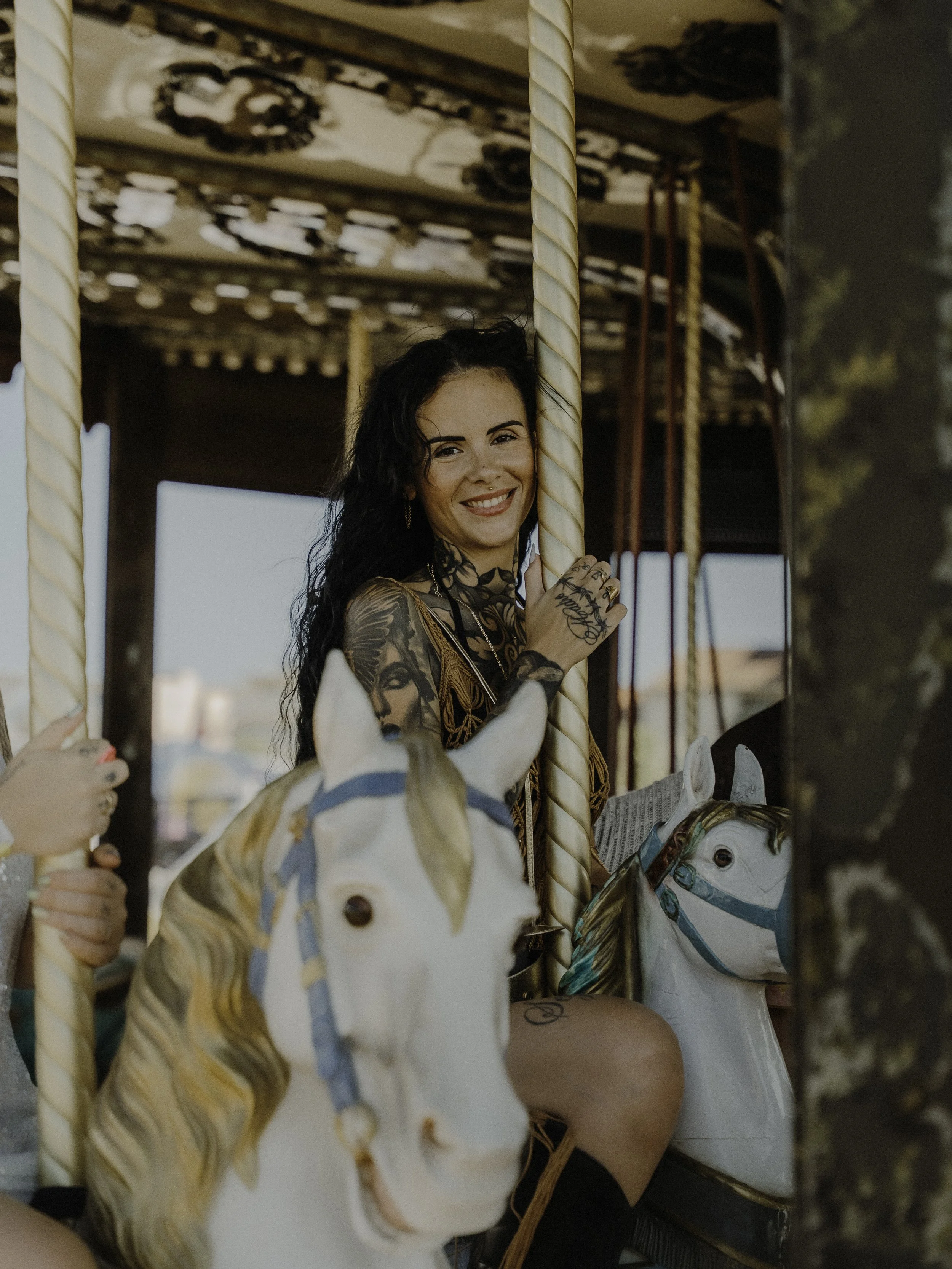 Jeune femme souriante avec tatouages, assise sur un manège à cheval en bois élégant, dans un parc d'attractions lors d'une journée ensoleillée.