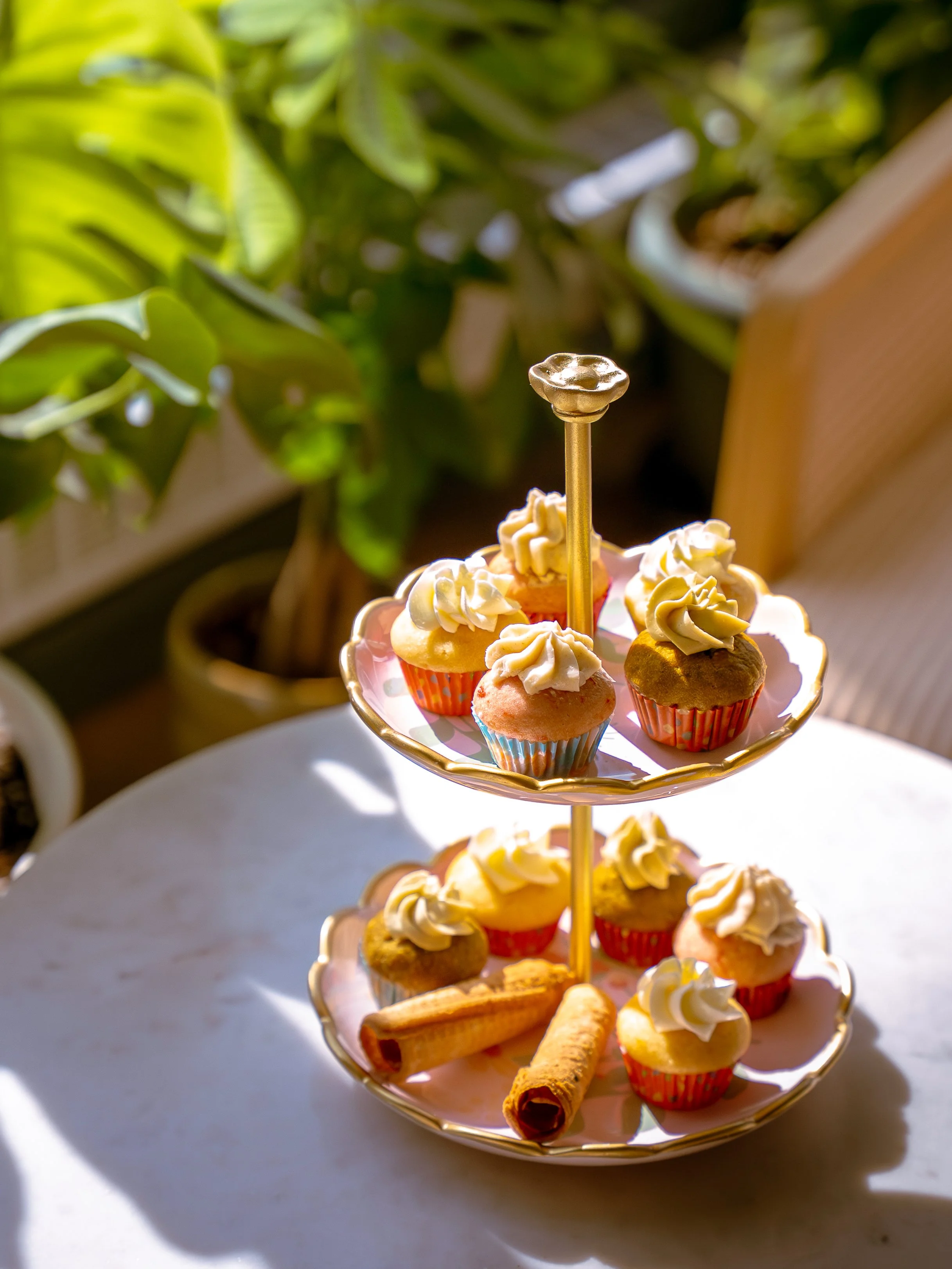 Plateau de cupcakes décorés avec de la crème, accompagné de biscuits roulés, sur une table en milieu intérieur avec des plantes en arrière-plan.