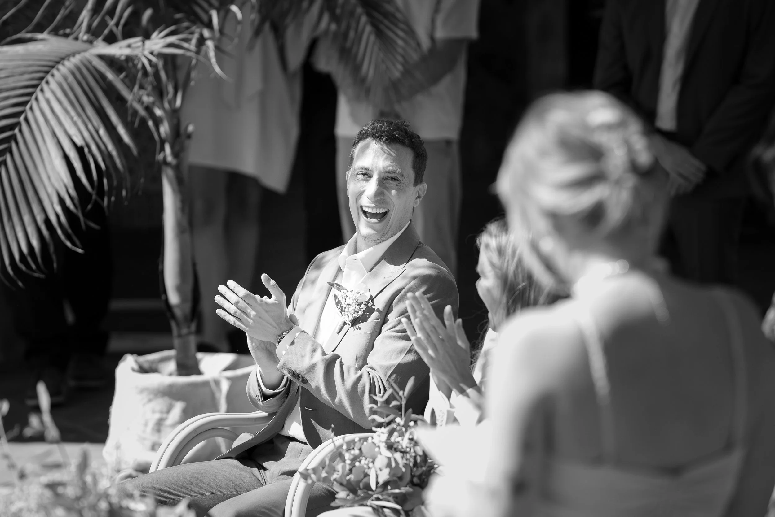 Un homme en costume souriant et applaudissant lors d'un mariage, entouré de personnes, en noir et blanc.
