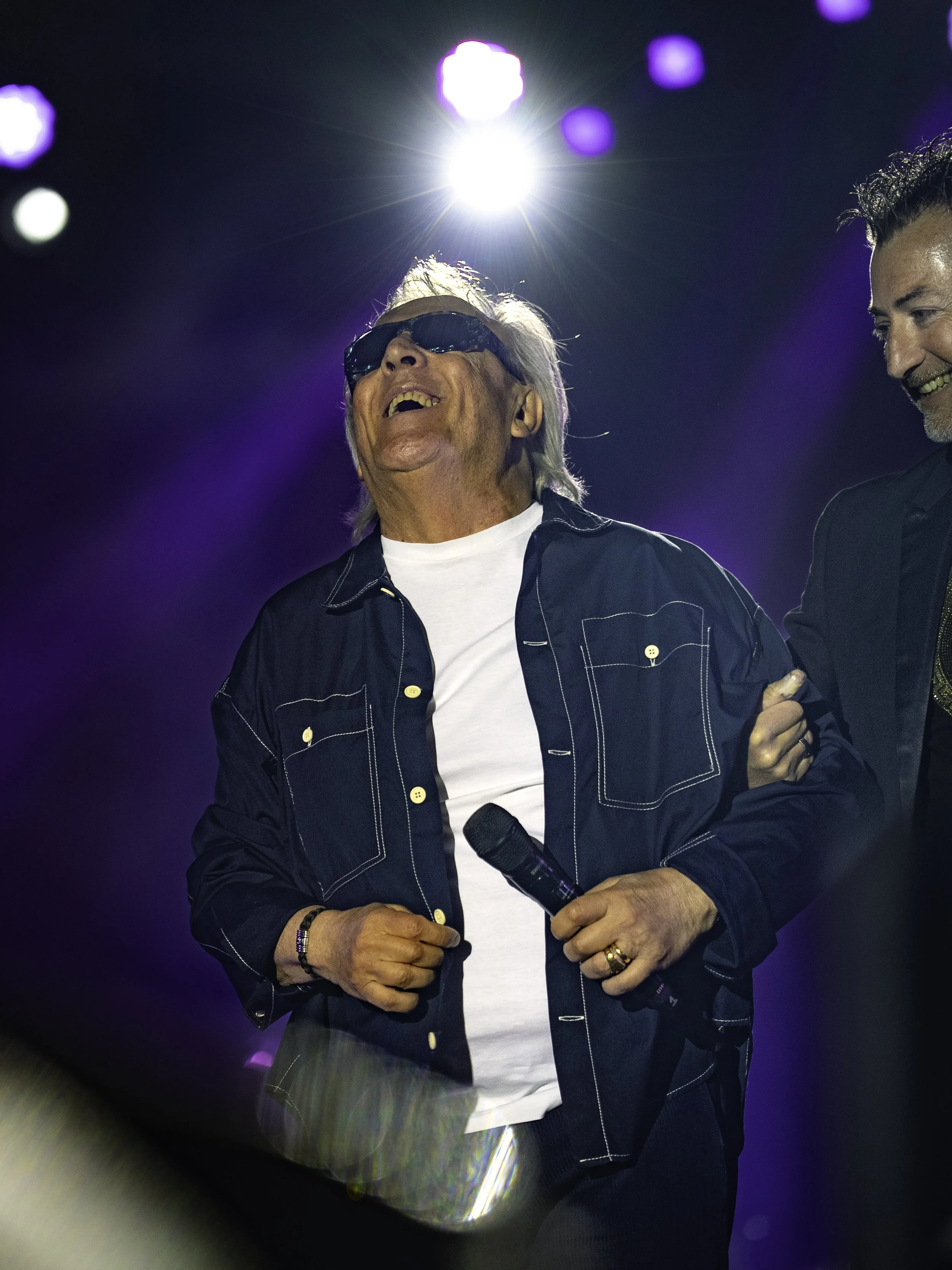 Un homme âgé portant des lunettes de soleil, une chemise noire à coutures blanches, une chemise blanche en dessous, tenant un micro dans une main, riant lors d'un concert ou d'un spectacle avec un éclairage coloré