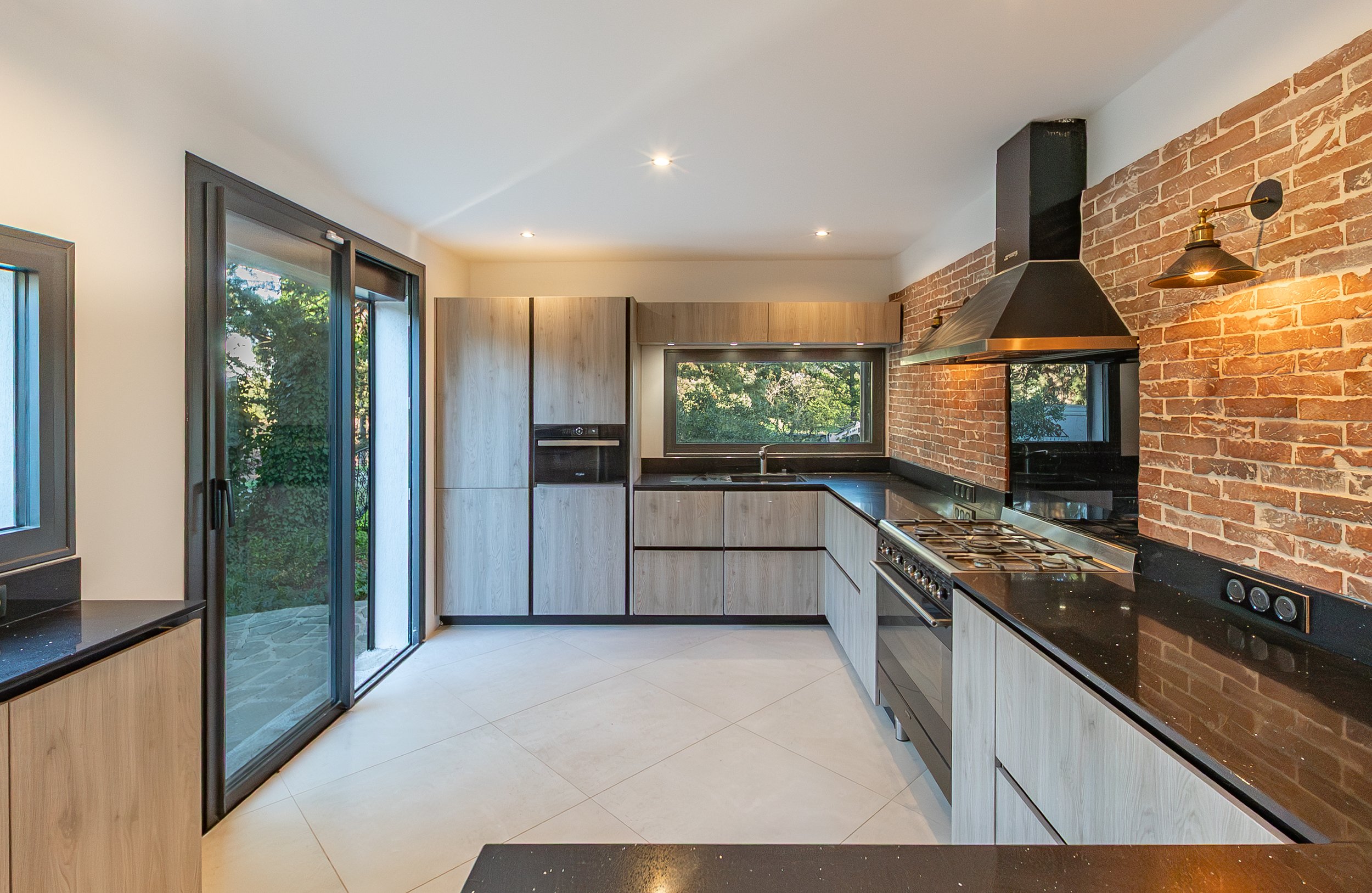 Cuisine moderne avec armoires en bois clair, mur en briques, plan de travail noir, fenêtre donnant sur l'extérieur, et porte-fenêtre en verre.