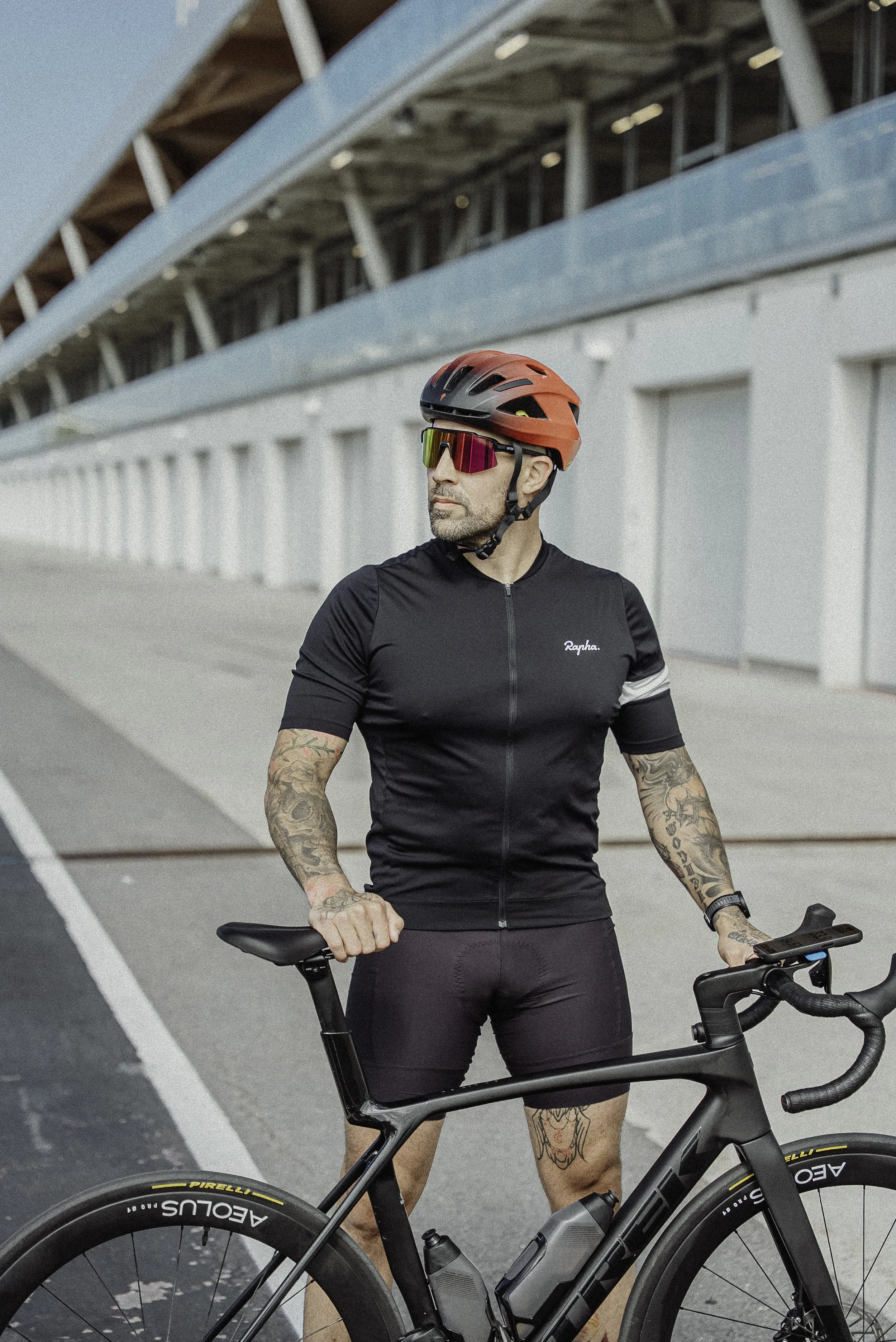 Un homme en tenue de cycliste avec un casque rouge et des lunettes de soleil, posant avec un vélo de route noir, devant un bâtiment moderne.