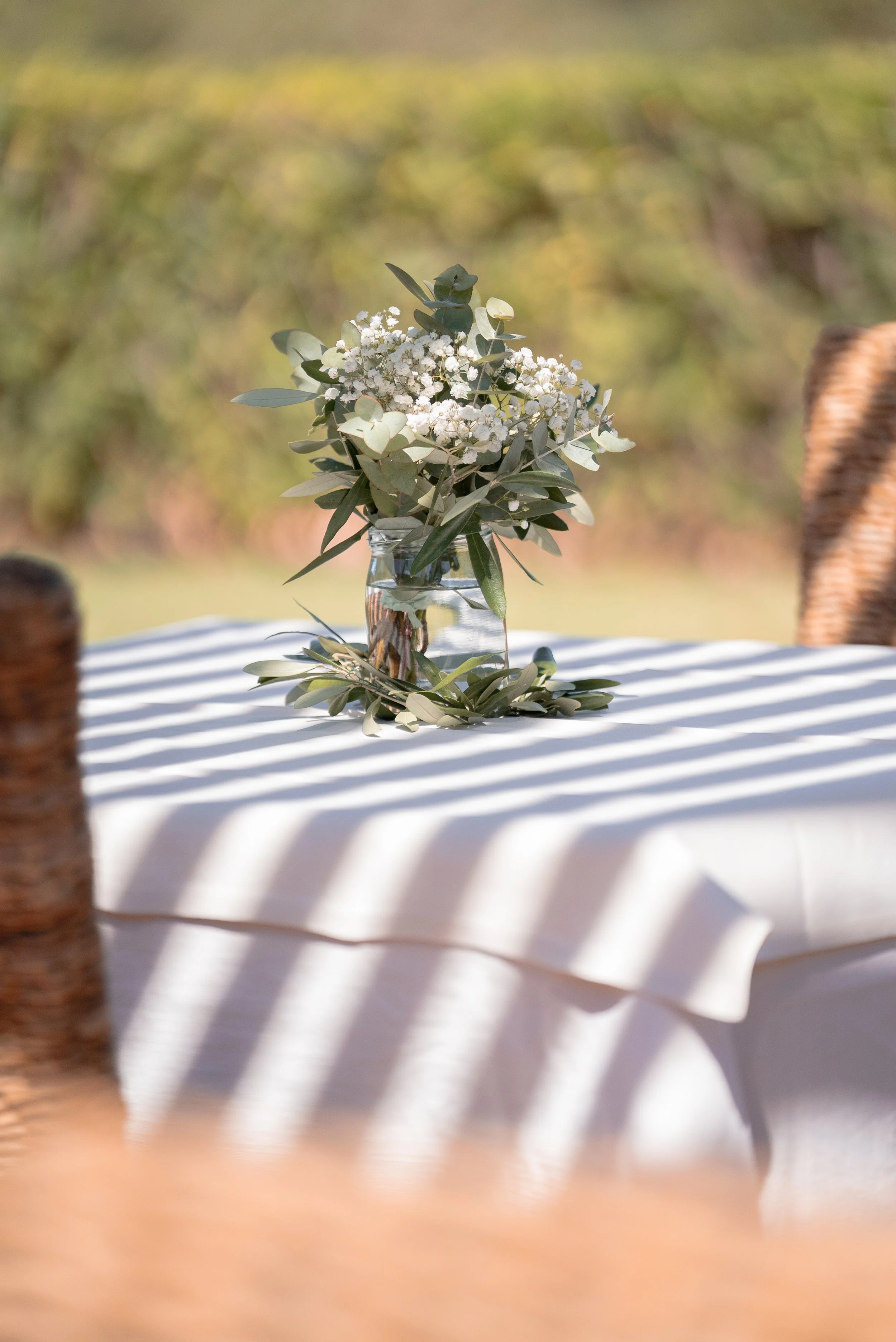 Un vase en verre avec des fleurs blanches et des feuilles vertes, placé sur une table recouverte d'une nappe blanche, avec des ombres créées par des bandes de lumière. En arrière-plan, un espace extérieur avec de la végétation verte floue.
