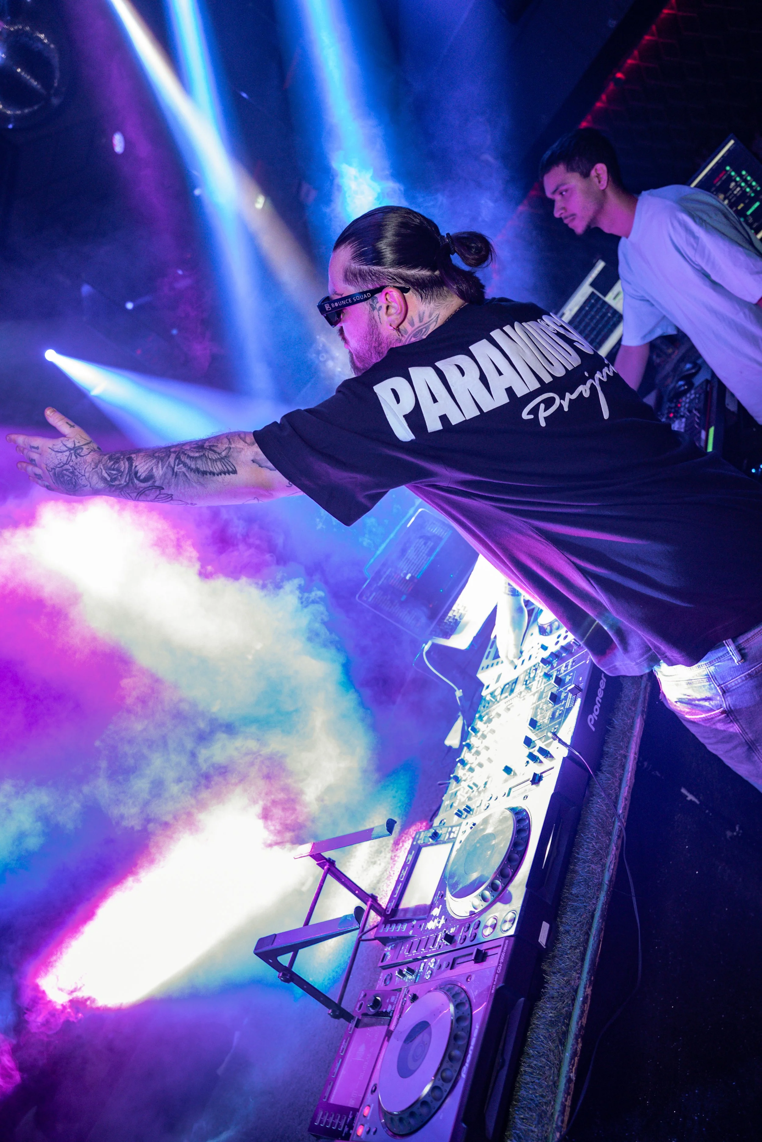 DJ avec tatouages et t-shirt « PARAMEDIC » en train de mixer sur scène avec des lumières colorées et de la fumée.