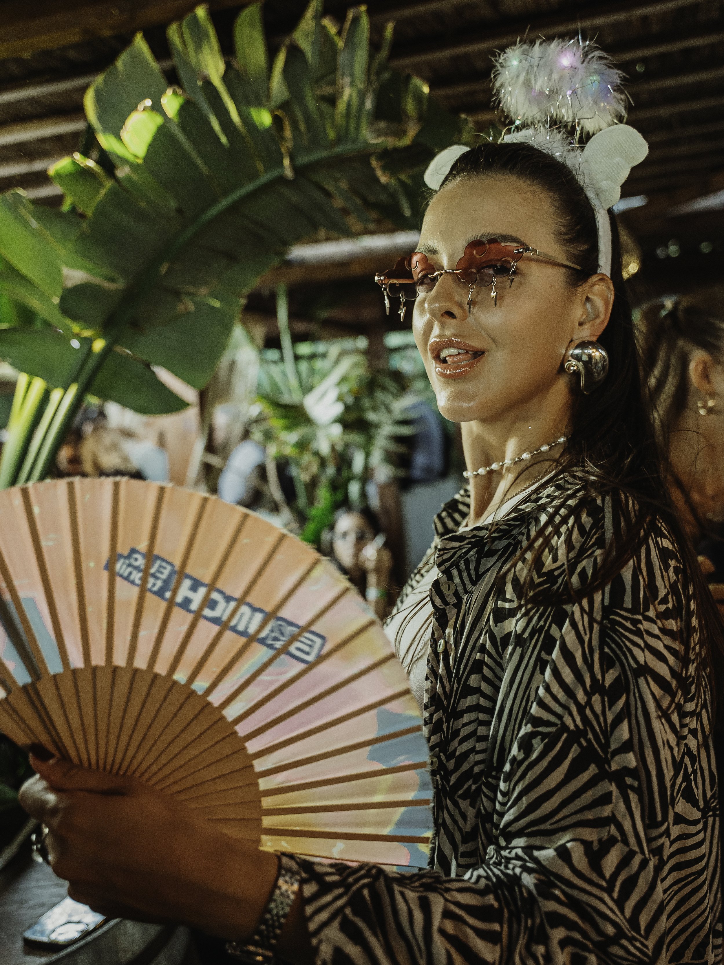 Une femme porte des accessoires inhabituels, des lunettes rouges avec des chaines et un headband avec des oreilles en peluche, elle tient un éventail en papier dans une ambiance de fête ou de marché.