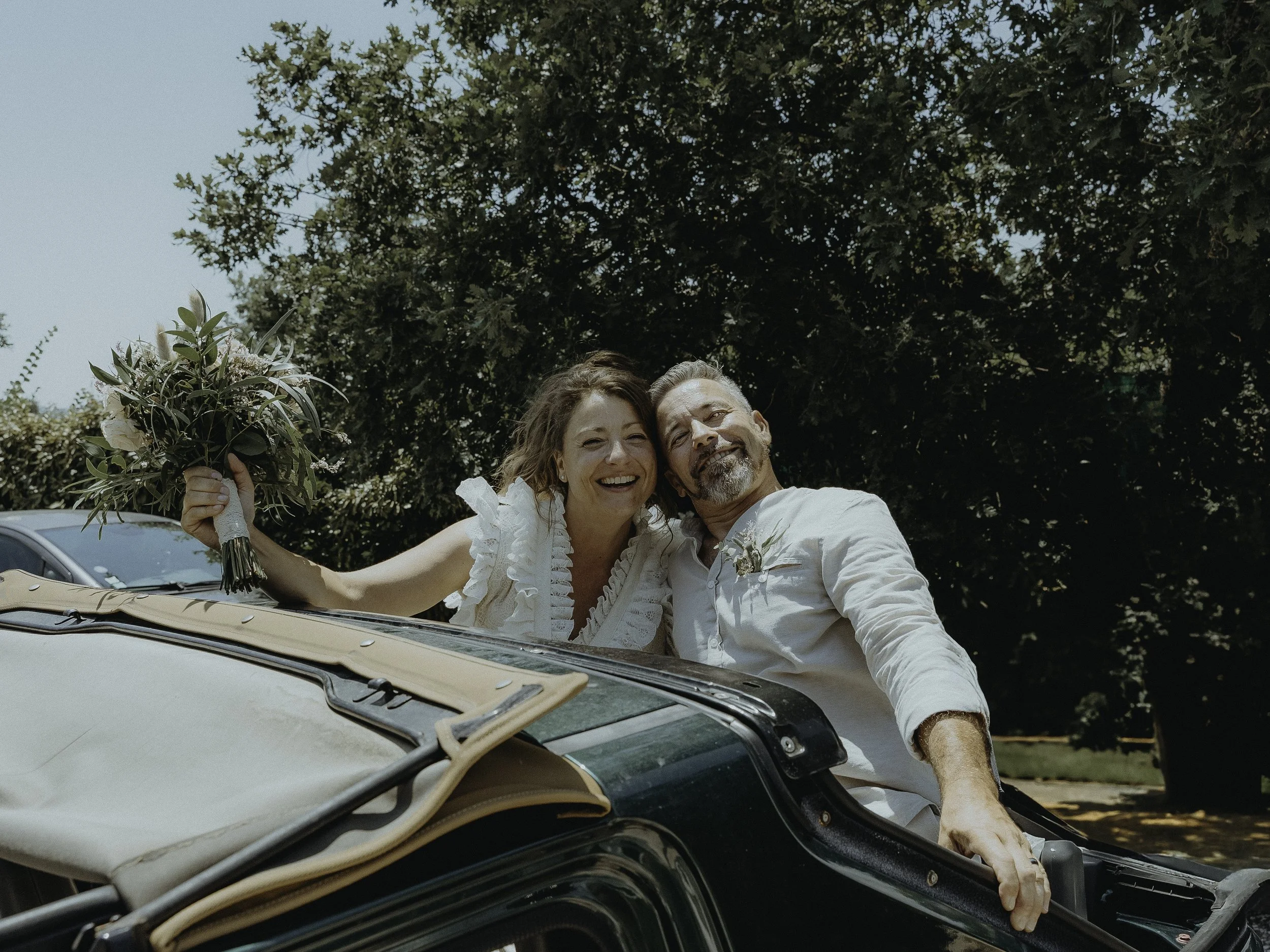 Un couple souriant, habillé en blanc, dans une voiture décapotable avec un bouquet de fleurs, lors d'une journée ensoleillée, entouré d'arbres.