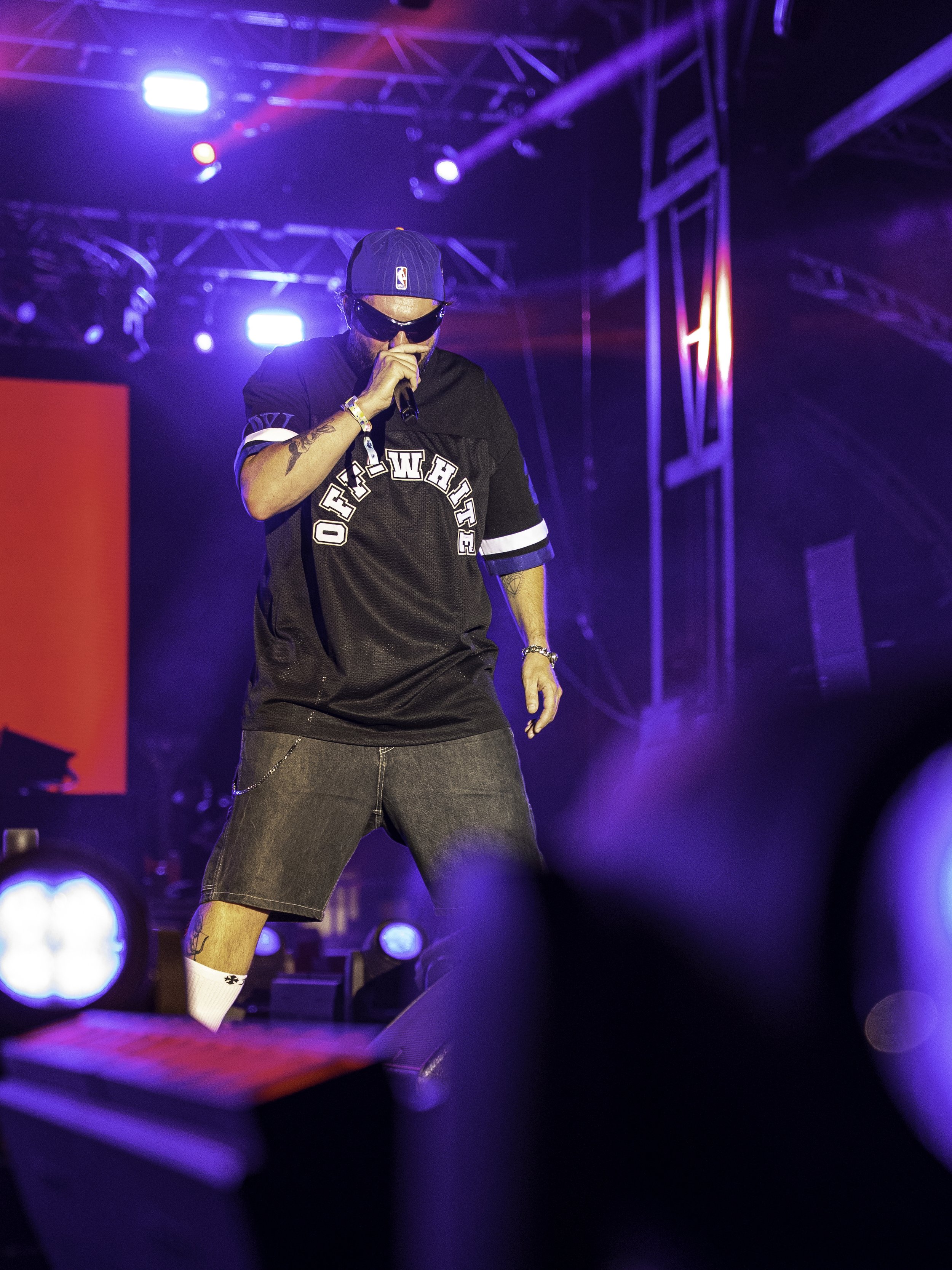 Un rappeur en tenue décontractée avec un t-shirt noir, un short en jean, une casquette et des lunettes de soleil, chante sur scène avec des lumières colorées en arrière-plan.