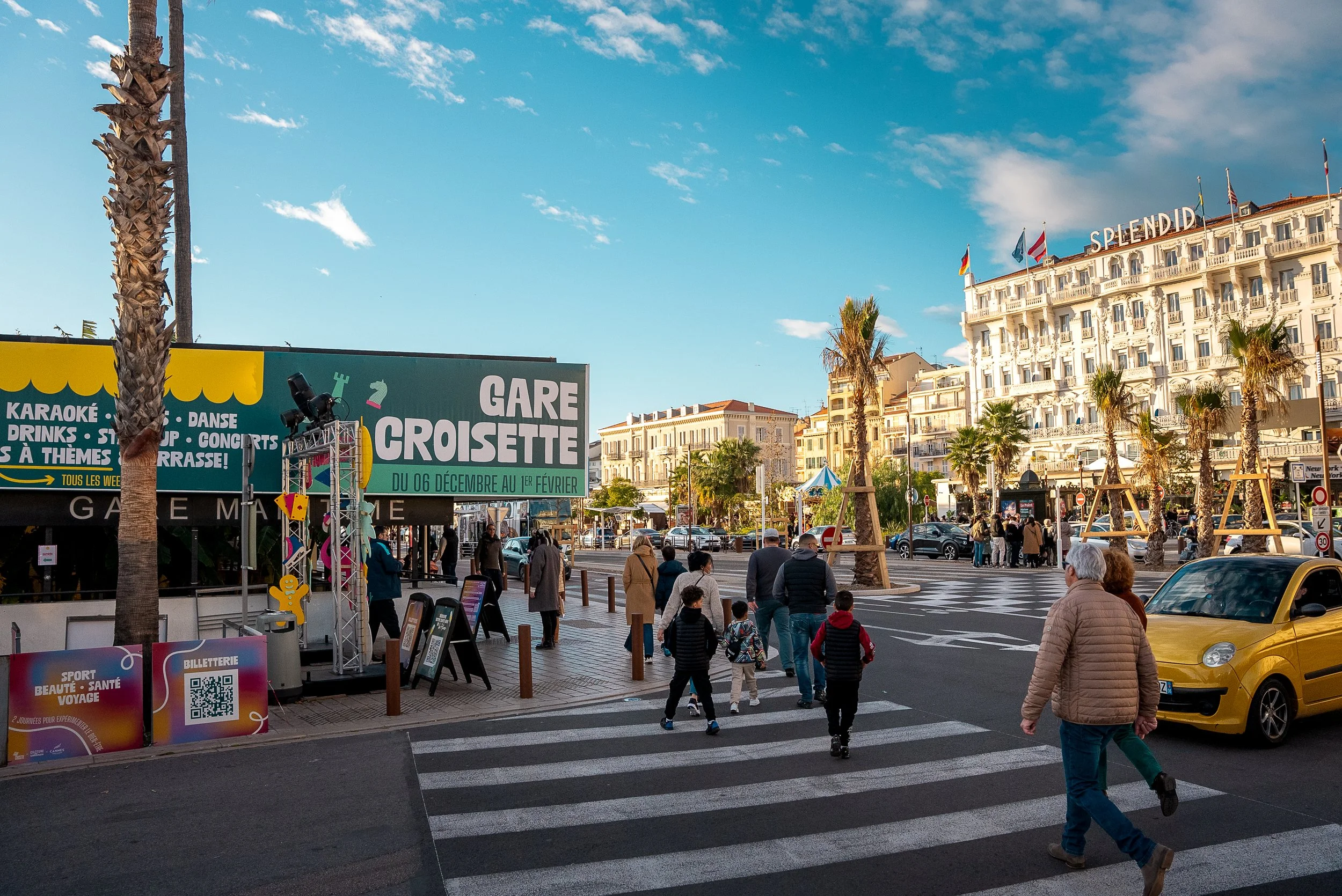 Vue d'une rue animée avec des personnes traversant un passage piéton, un grand hôtel blanc portant le nom 'Splendid', palmier, voitures jaunes et un panneau publicitaire pour un événement de karaoké et concerts.