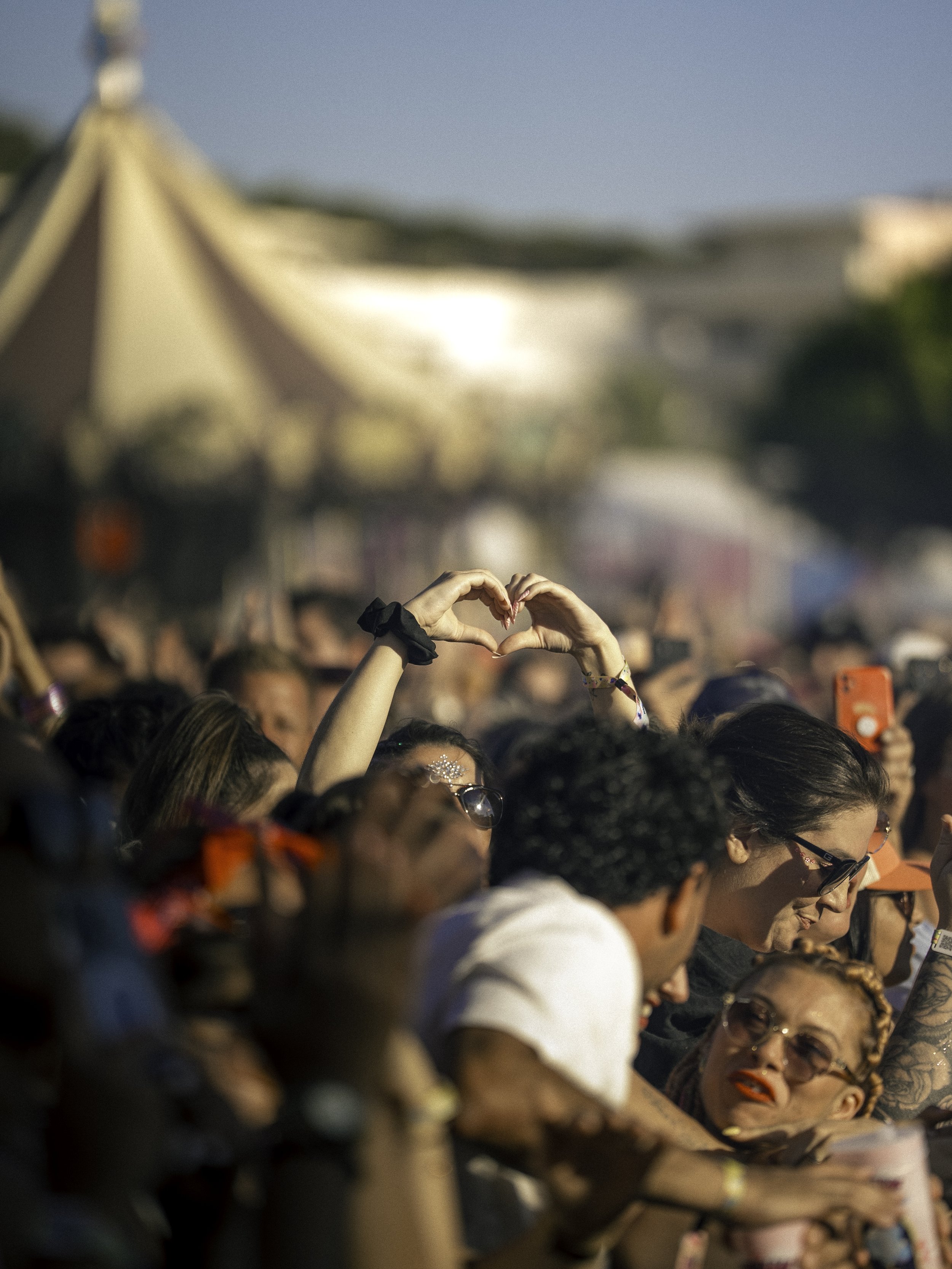 Une foule de personnes assistent à un événement en plein air, avec une personne formant un cœur avec ses mains, en arrière-plan un chapiteau.