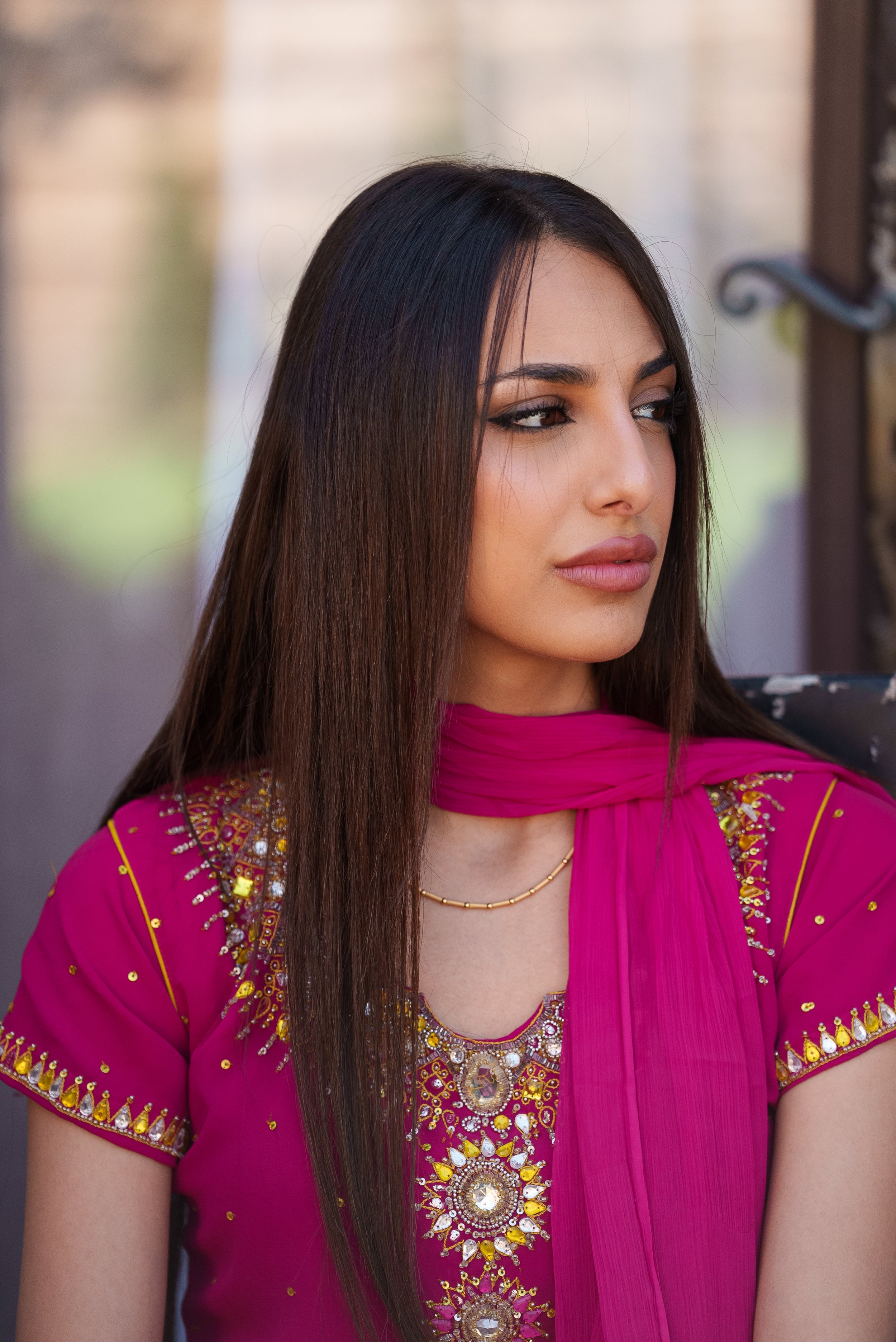 Une femme portant une tenue traditionnelle rose foncé avec des détails dorés et des pierres précieuses, assise dehors, regardant sur le côté.
