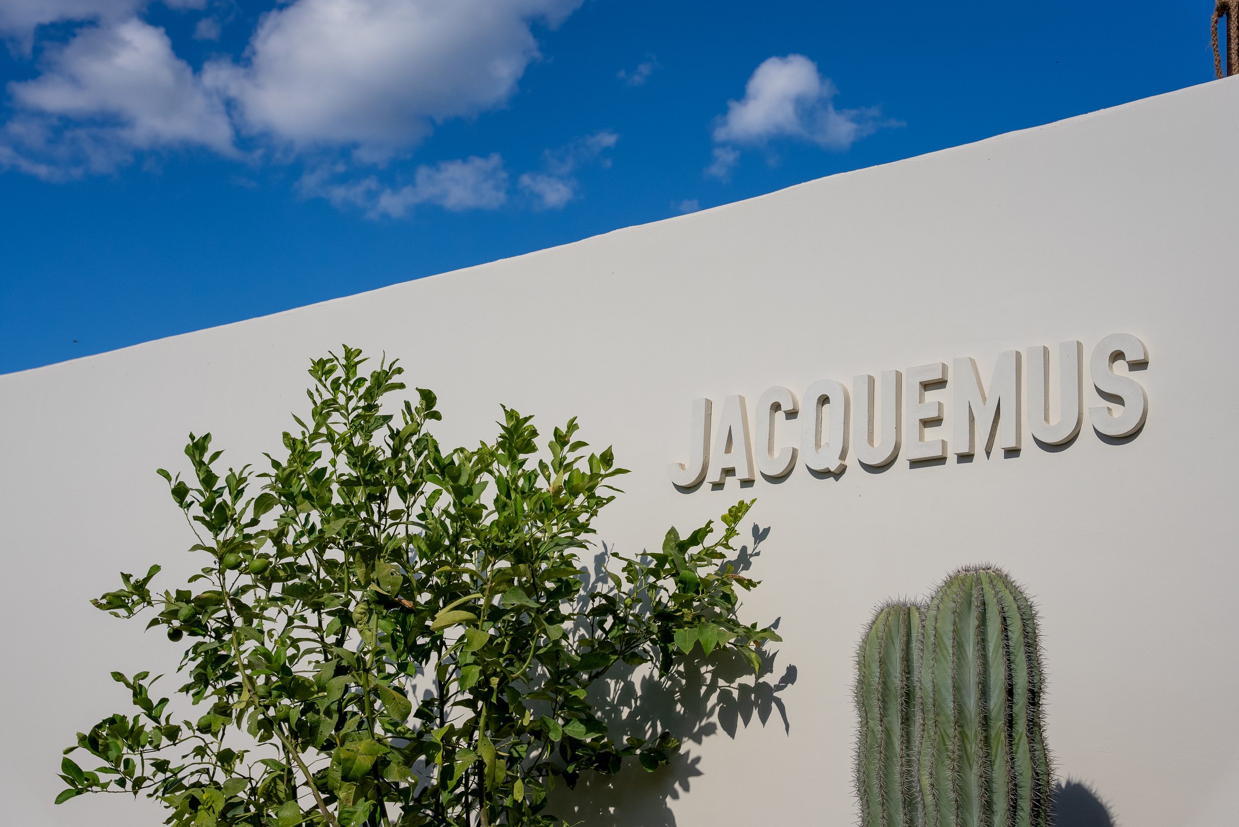 Une façade blanche avec le nom 'JACQUEMUS', des plantes dont un cactus, et un ciel bleu avec quelques nuages.