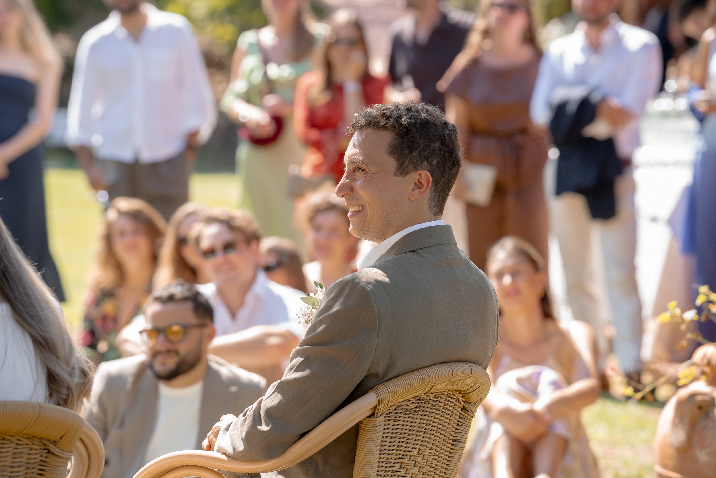 Un homme souriant en costume beige lors d'un mariage en extérieur, entouré de plusieurs personnes assises ou debout.