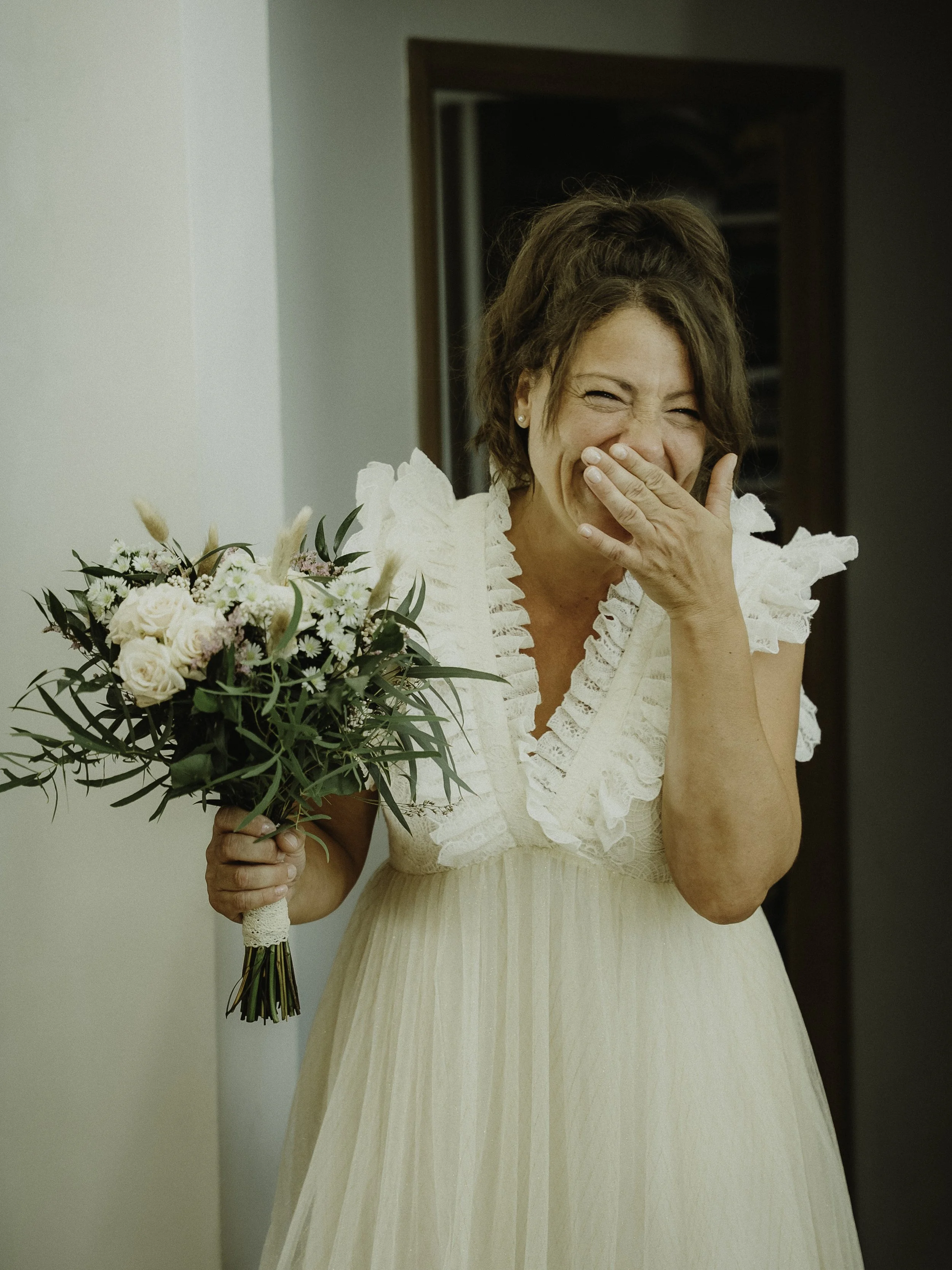 Une femme souriante en robe blanche tient un bouquet de fleurs et se cache la bouche avec la main.