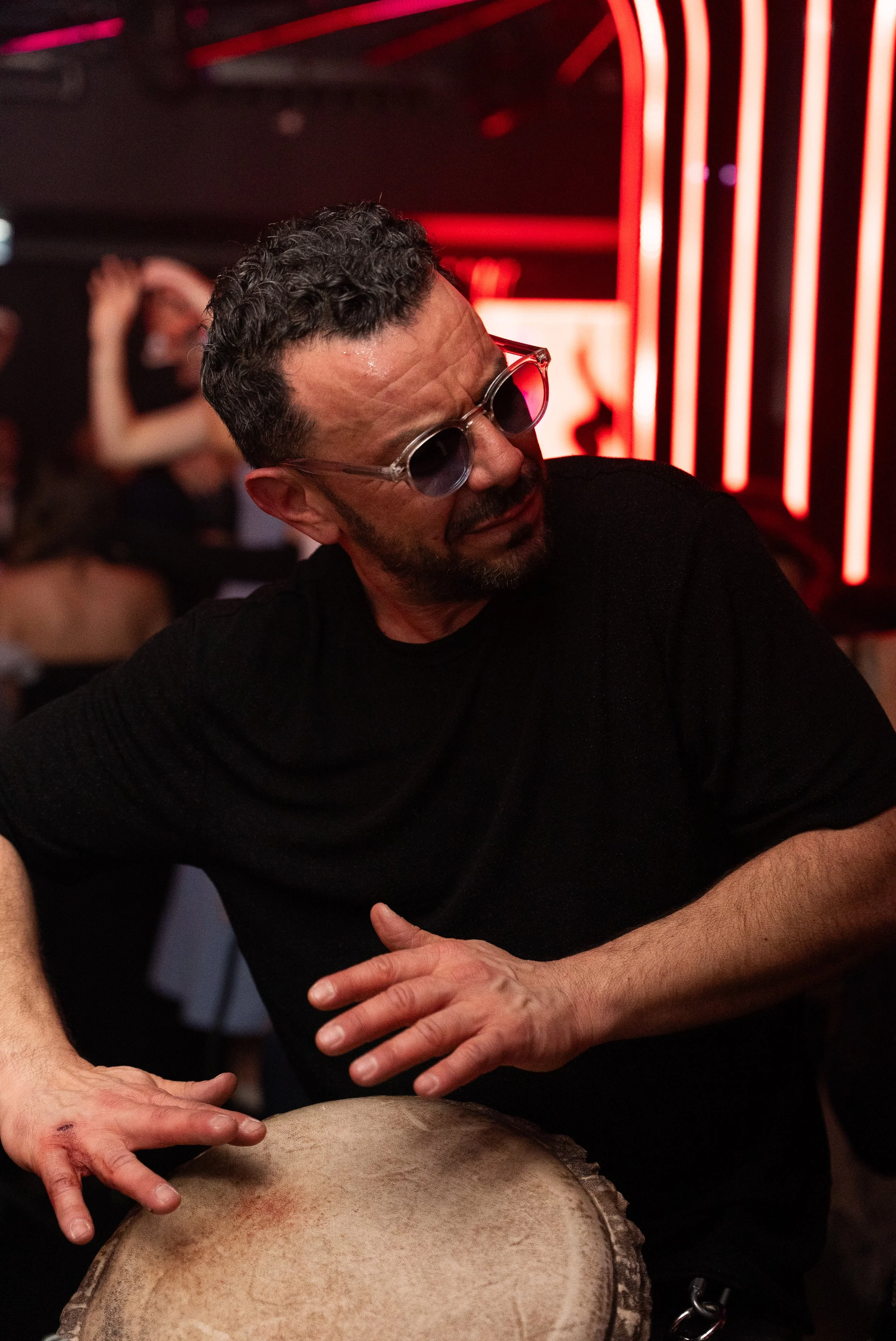 Un homme avec des lunettes rondes joue du tambour dans un environnement sombre avec des lumières rouges.