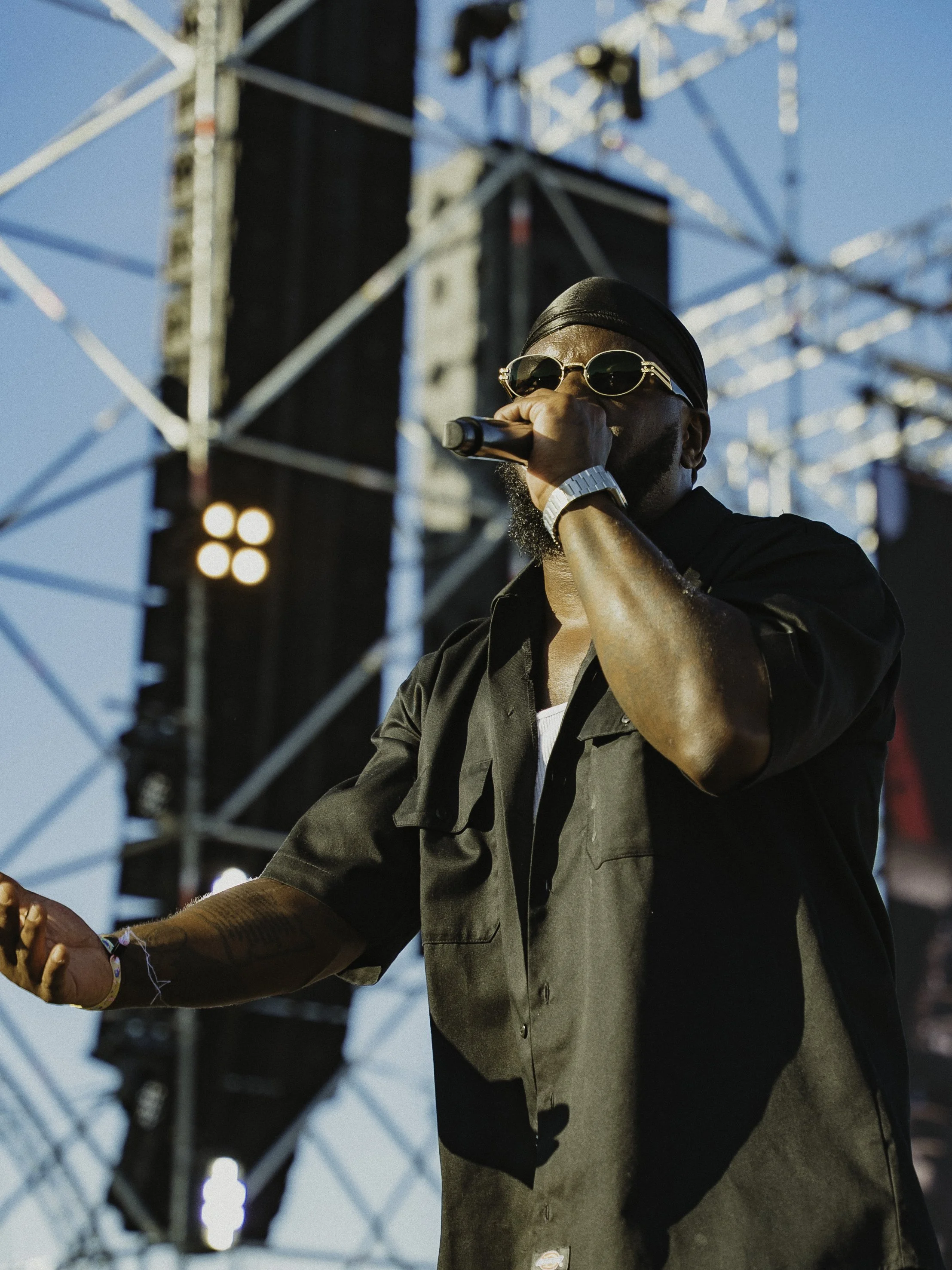 Un homme portant des lunettes de soleil, un bandeau noir et un bracelet en train de chanter ou parler dans un microphone lors d'un concert en plein air, avec une scène et une structure métallique en arrière-plan.