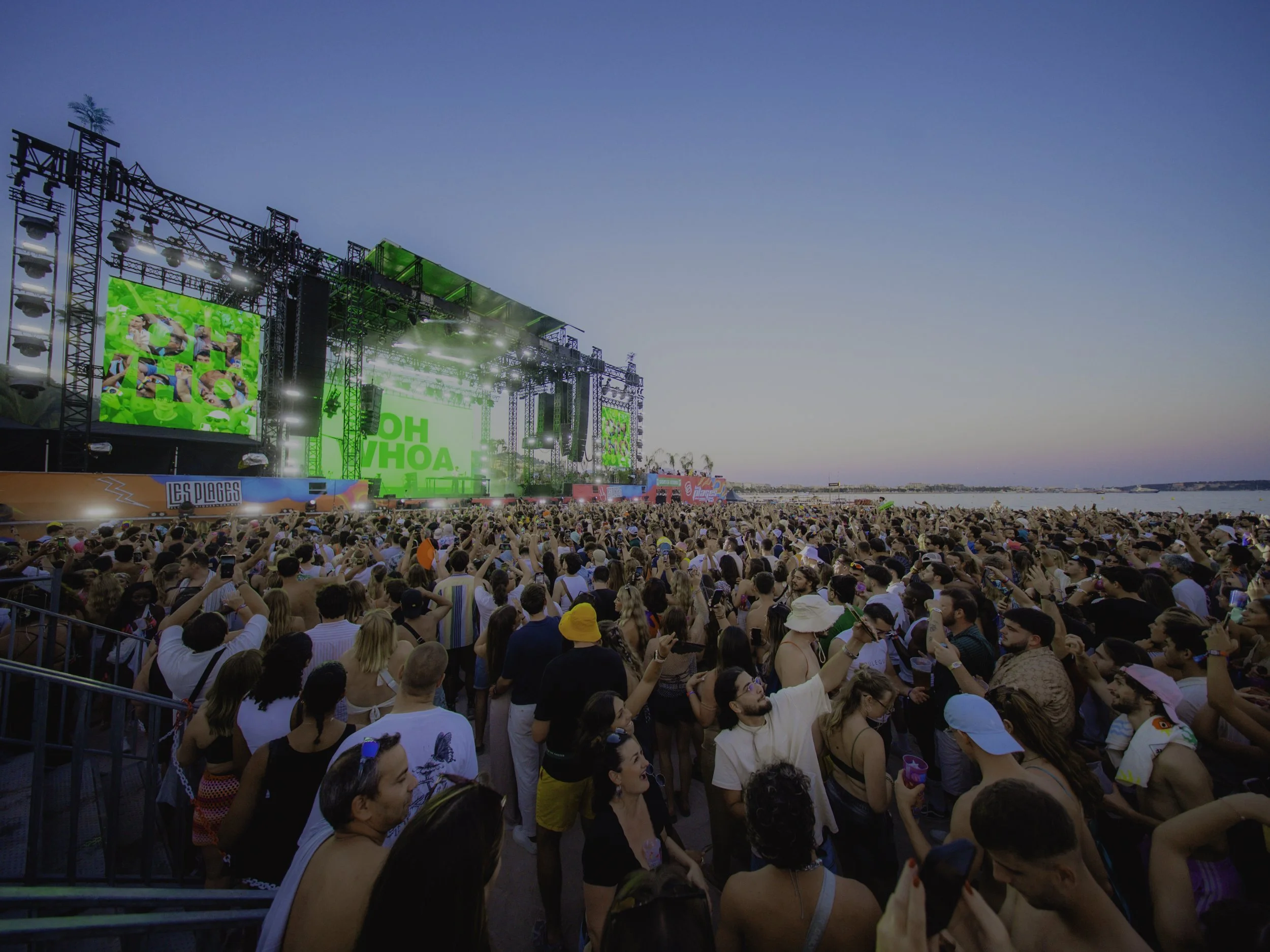 Un grand public assiste à un concert en plein air près de la mer au coucher du soleil, avec une scène lumineuse et des écrans géants.