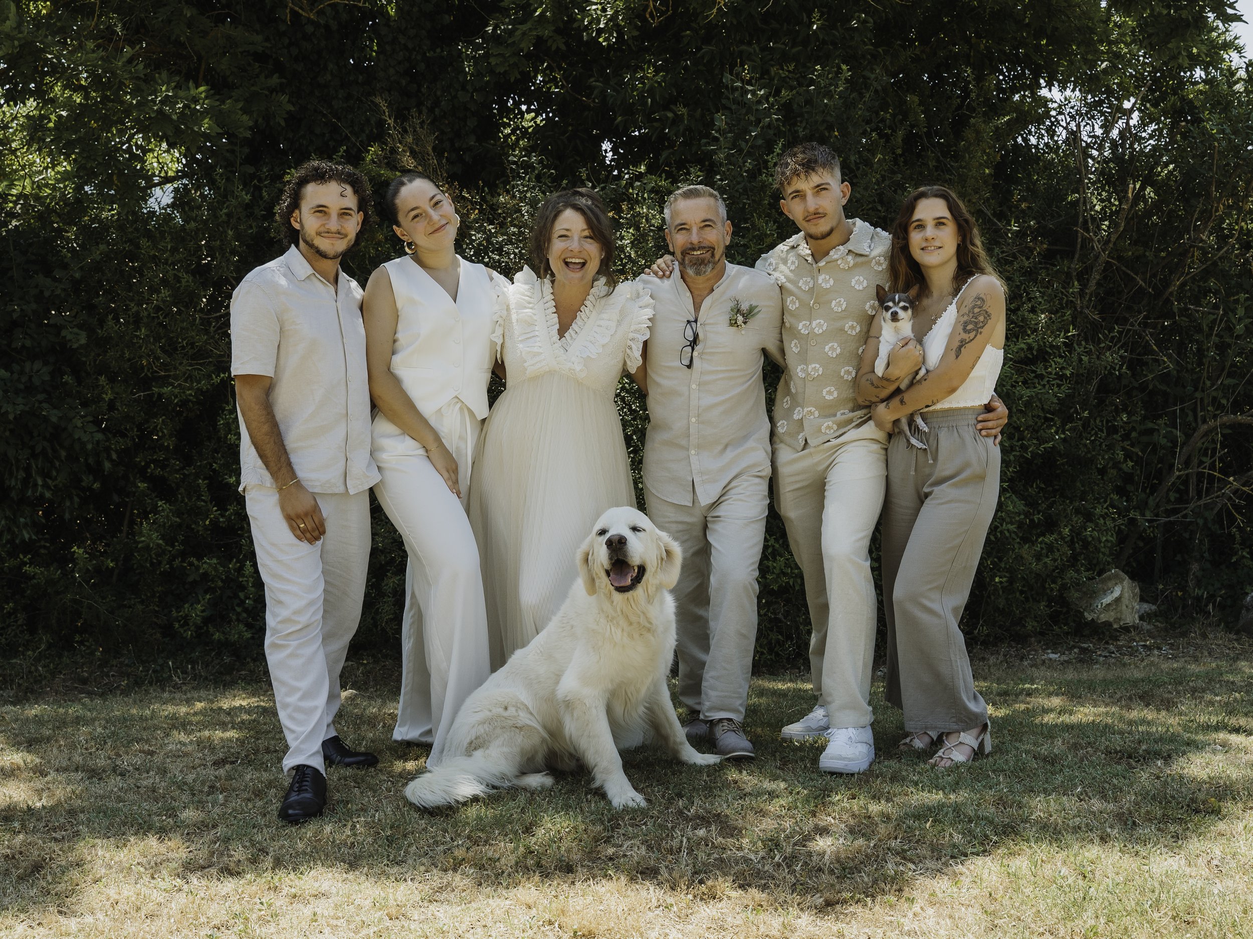 Groupe de sept personnes souriantes avec deux chiens, réunies en extérieur dans un cadre verdoyant, lors d'une occasion spéciale ou d'une célébration.