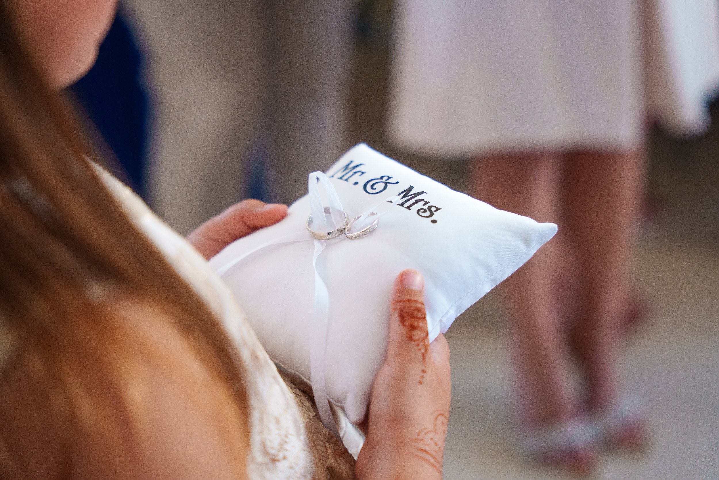 Une femme tient un coussin blanc avec la inscription 'Mr. & Mrs.' et deux alliances en satin blanc enroulées autour d'un ruban, lors d'une cérémonie de mariage.