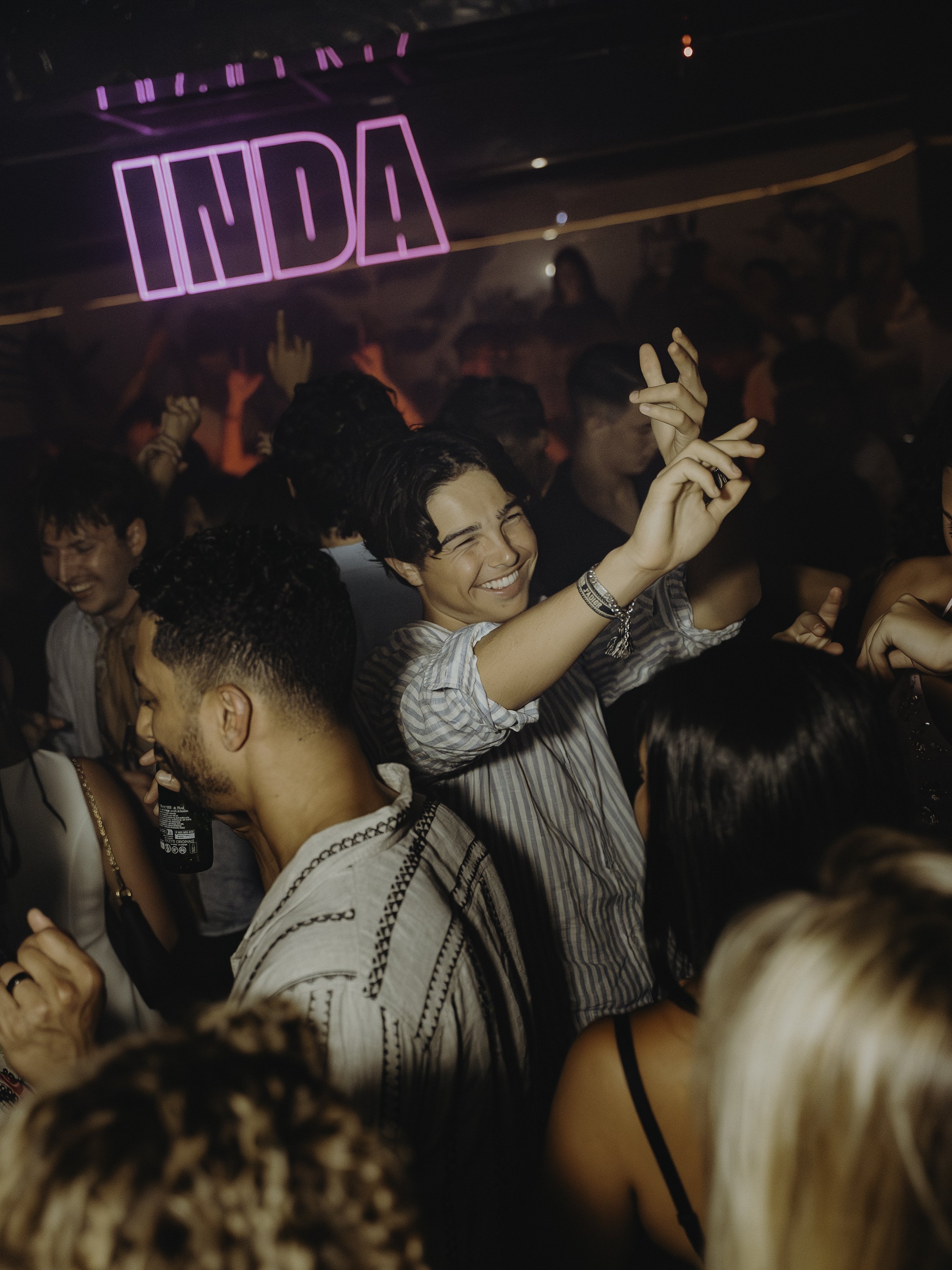 Groupe de personnes dansant et s'amusant dans une fête en intérieur, avec une enseigne lumineuse rose en arrière-plan qui dit 'LIDAS'.
