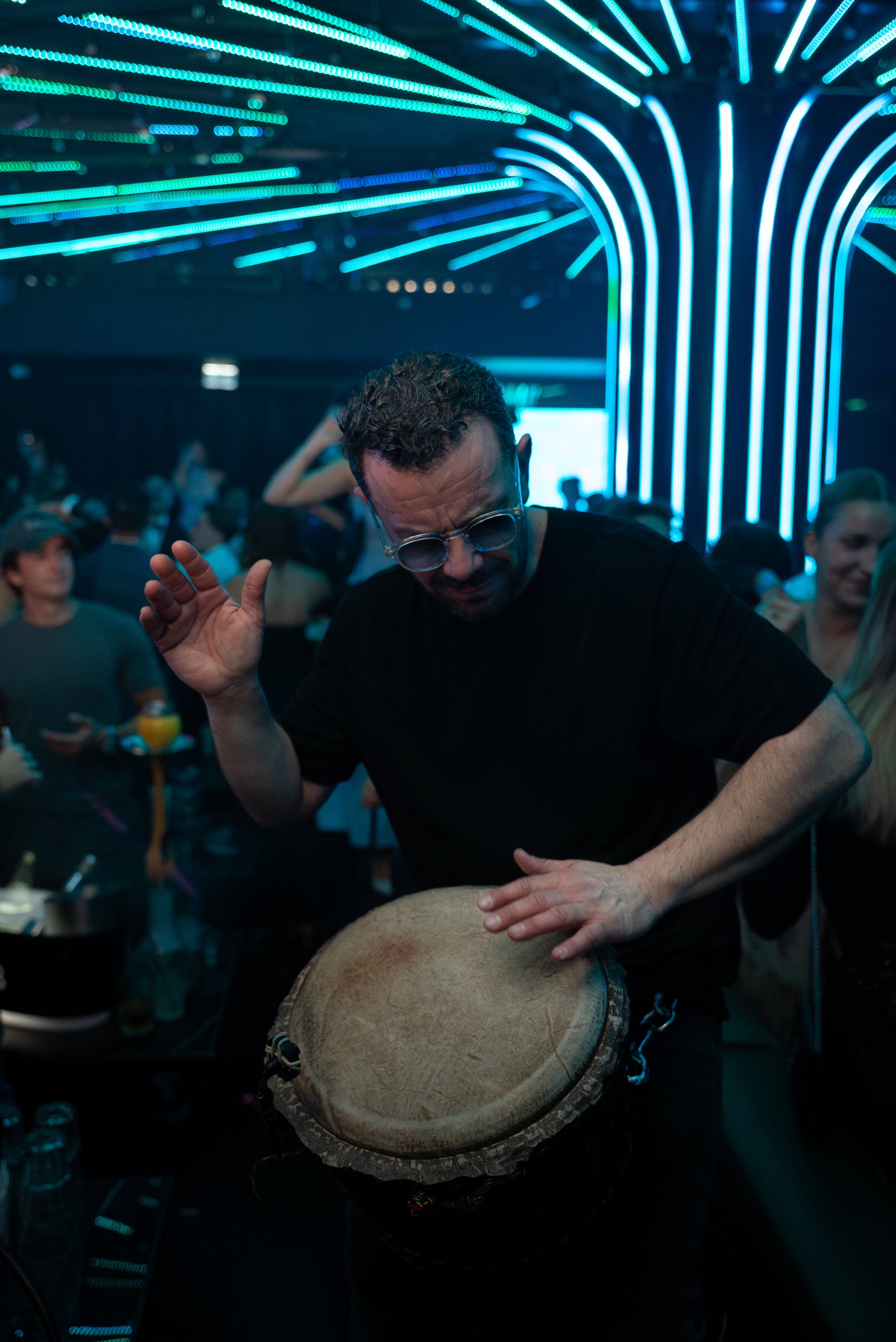Un homme jouant du djembé dans une ambiance de fête avec un plafond illuminé de lumière bleue et des personnes dansant et regardant autour.
