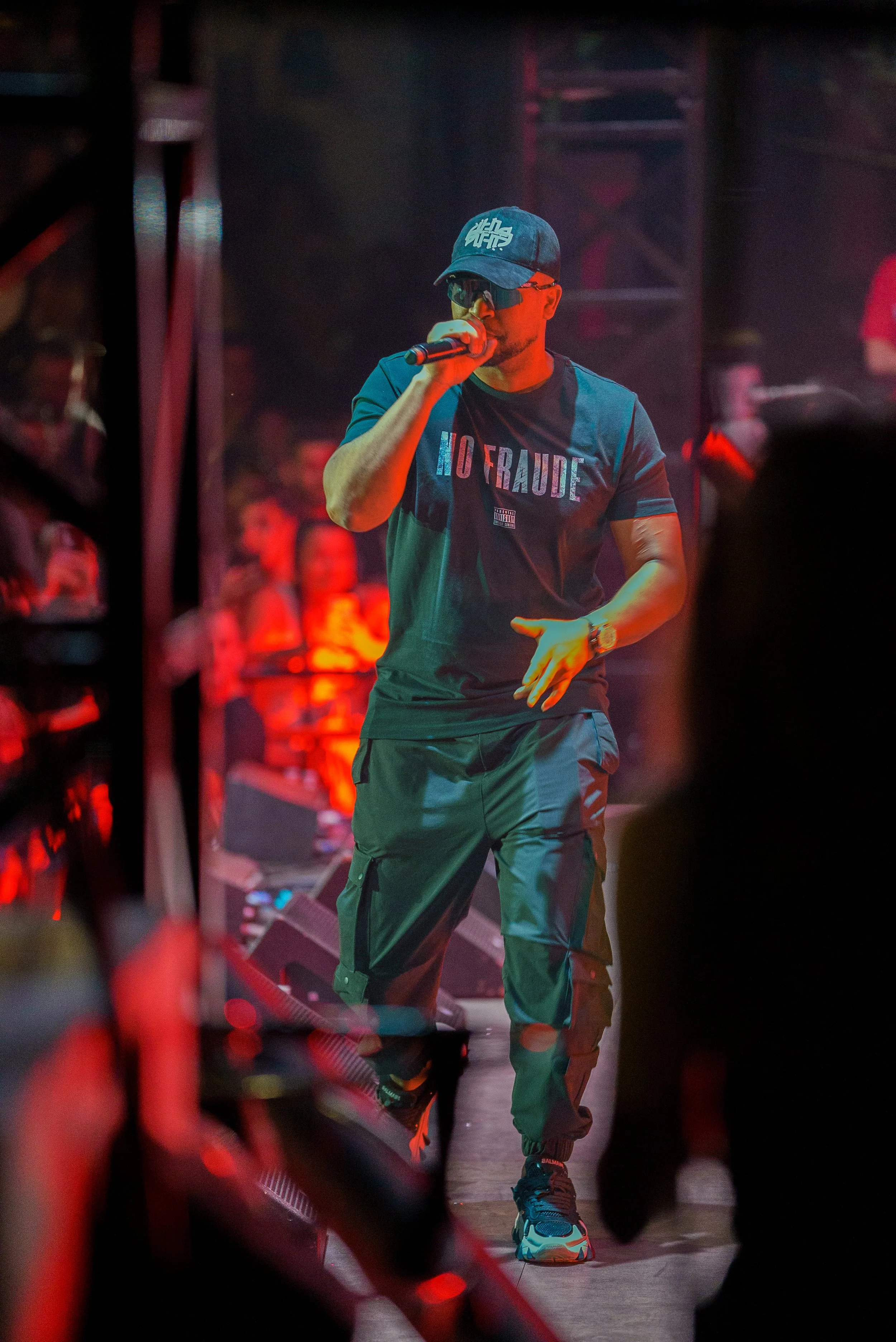 Un homme portant une casquette, des lunettes de soleil et un t-shirt avec le texte 'NO FRAUDE' chante ou parle dans un micro lors d'une performance en intérieur, avec un public en arrière-plan.