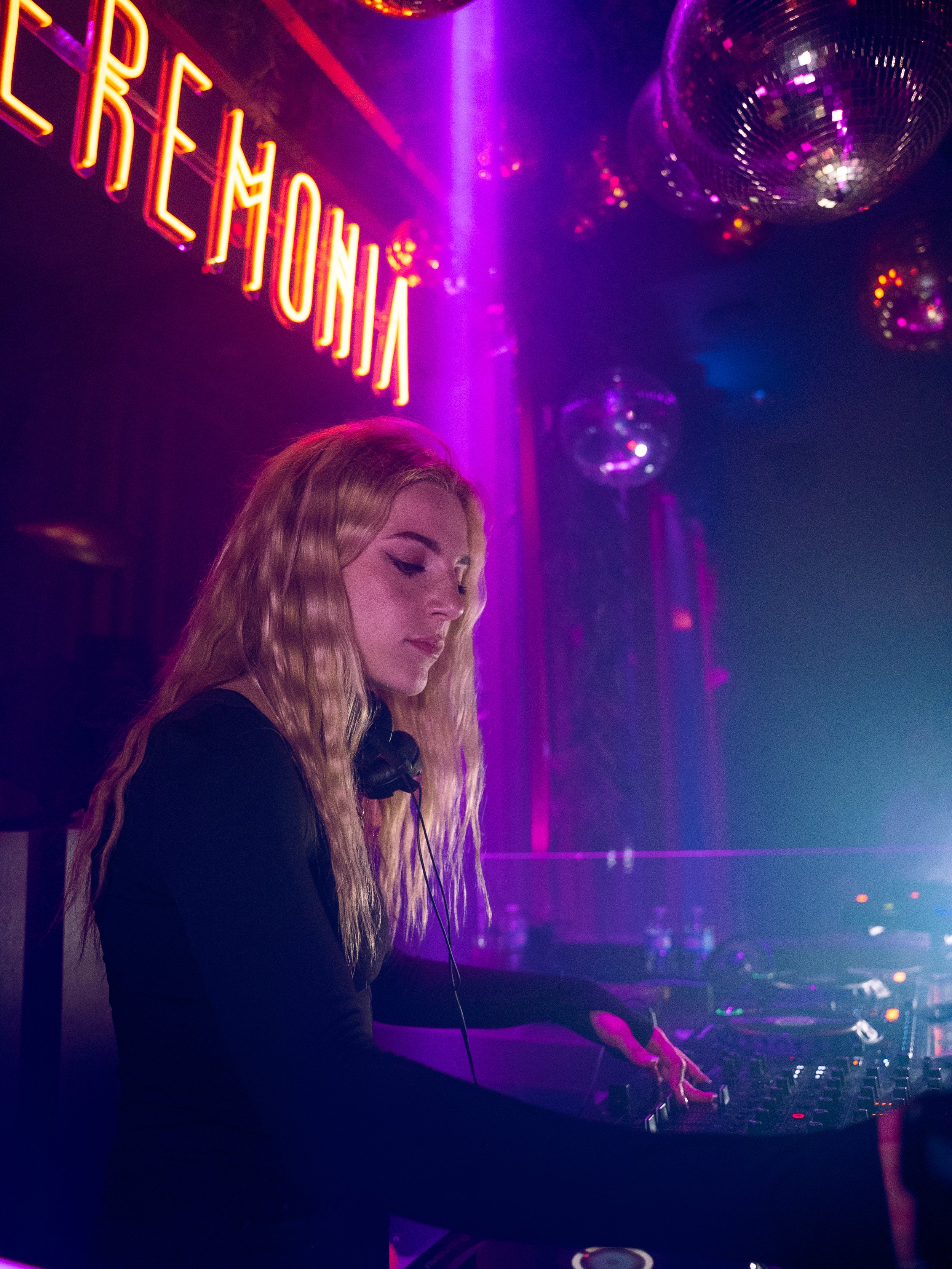 Une jeune femme DJ mixant dans un club avec des lumières violettes et des boules disco au plafond.
