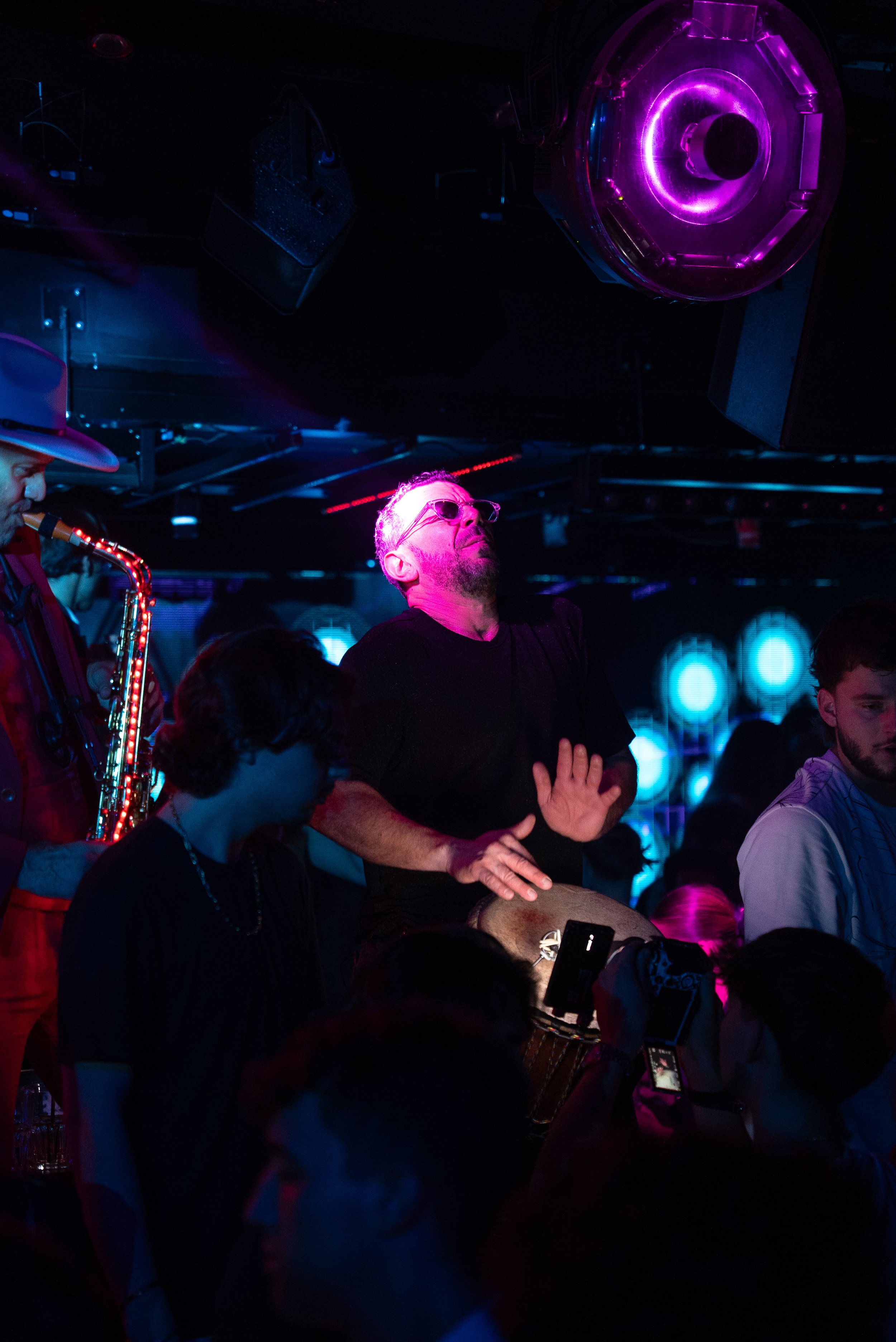 Un groupe de personnes dans une ambiance de club ou concert, avec des lumières néon violettes et bleues, une personne jouant du saxophone avec un chapeau, un homme en lunettes et noir qui semble profiter de la musique, et d'autres personnes qui regar