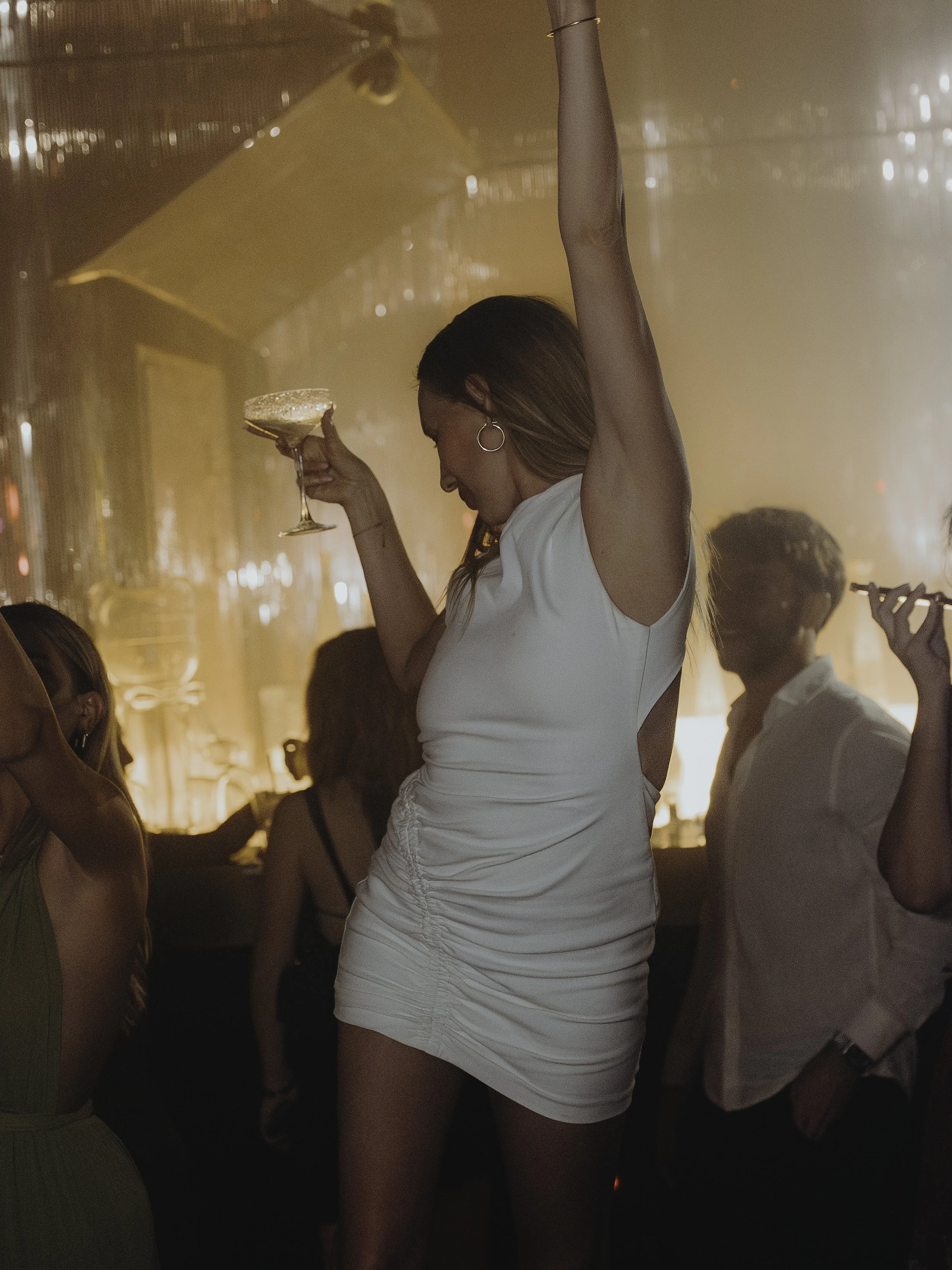 Une femme danse dans une boîte de nuit, portant une robe courte blanche, tenant un verre à cocktail, avec plusieurs autres personnes dans l'arrière-plan.