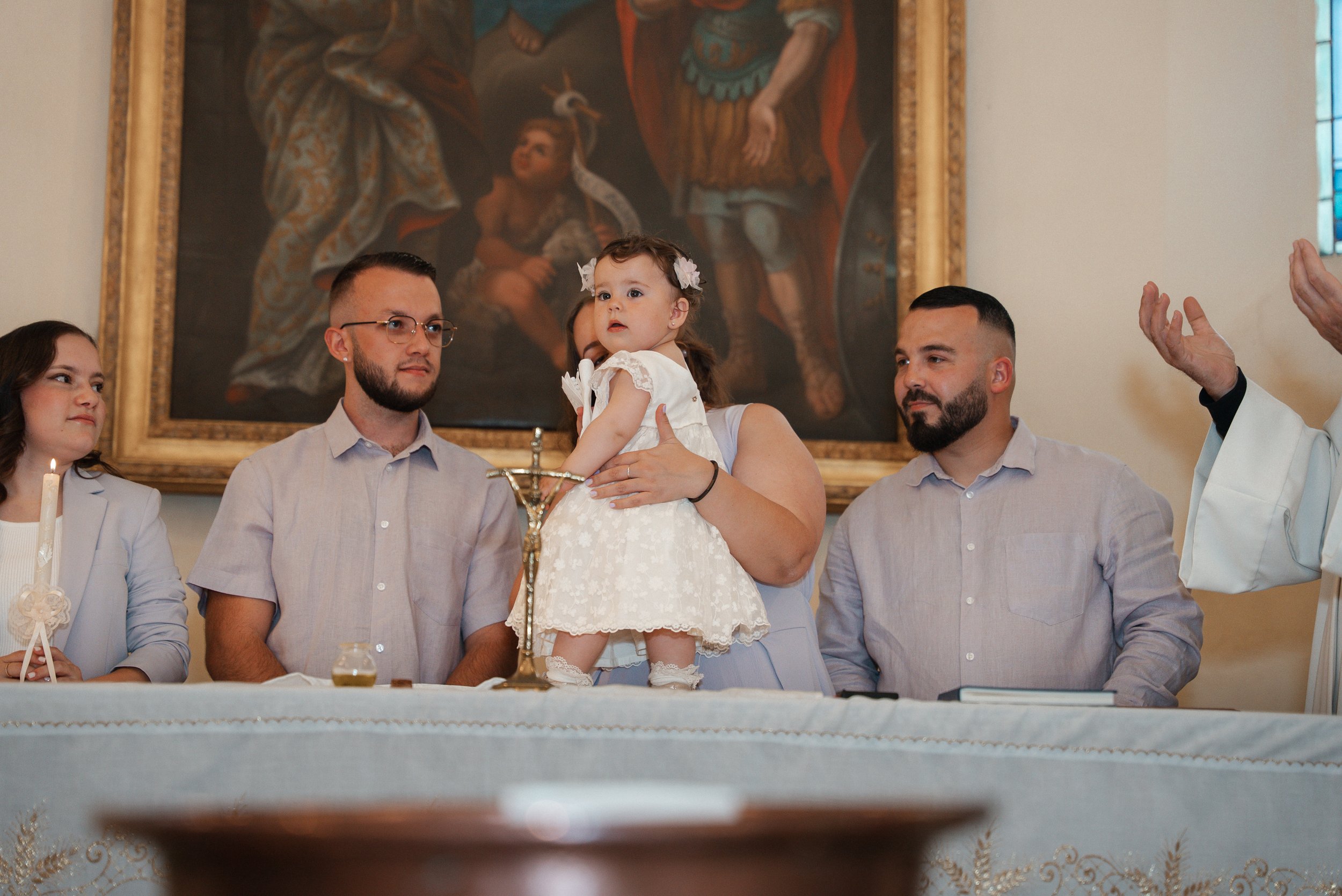 Une cérémonie religieuse avec une petite fille en robe blanche au centre, entourée de quatre adultes assis à une table, dans une église, avec un tableau religieux en arrière-plan.