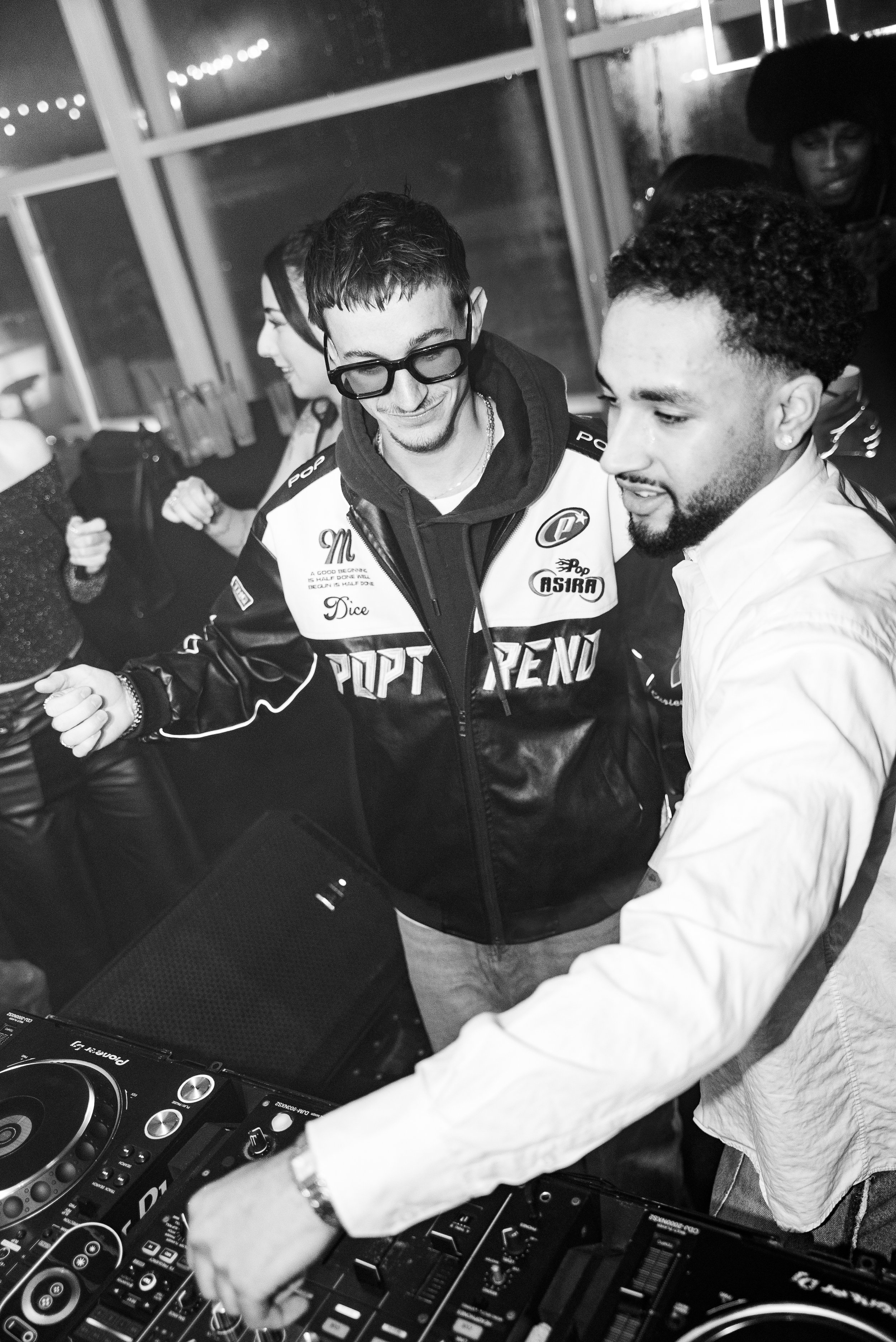 Deux hommes derrière un DJ, un d'eux ajuste la musique, souriant dans un cadre intérieur occupé par plusieurs autres personnes, en noir et blanc.