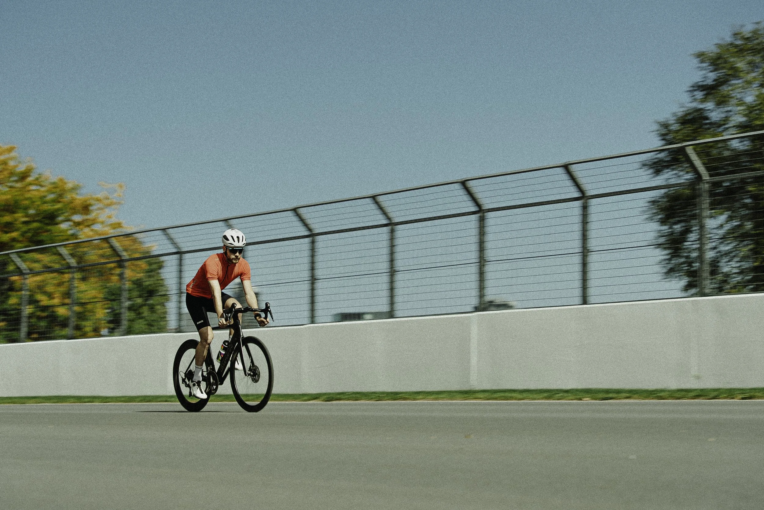 Un homme en vélo, portant un casque blanc et une tenue rouge, roulat sur une piste au soleil avec un ciel bleu et des arbres en arrière-plan.