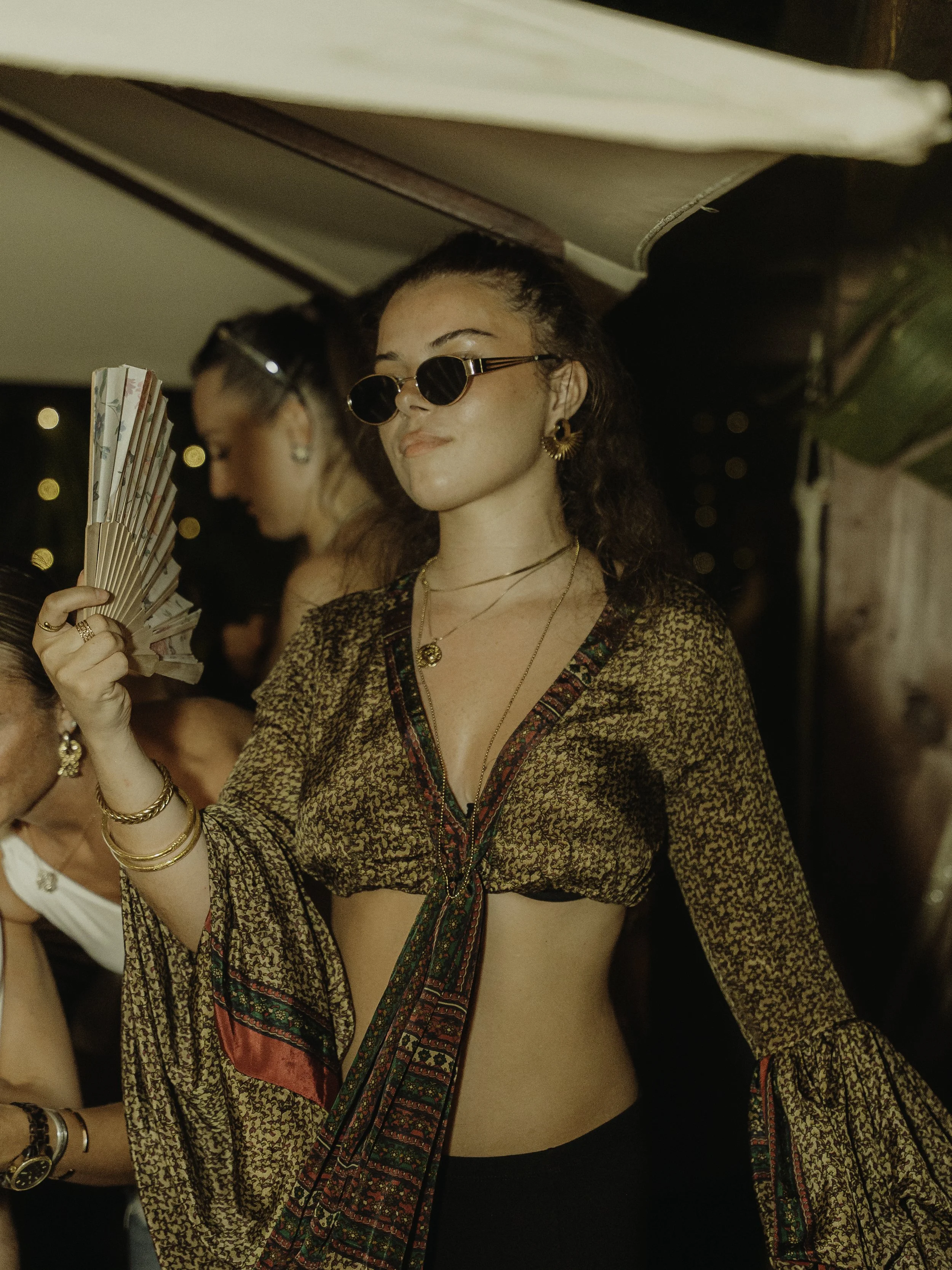 Une femme porte des lunettes de soleil rondes, des bijoux dorés, et un haut à manches longues avec un foulard coloré attaché autour de sa taille dans un environnement festif la nuit.