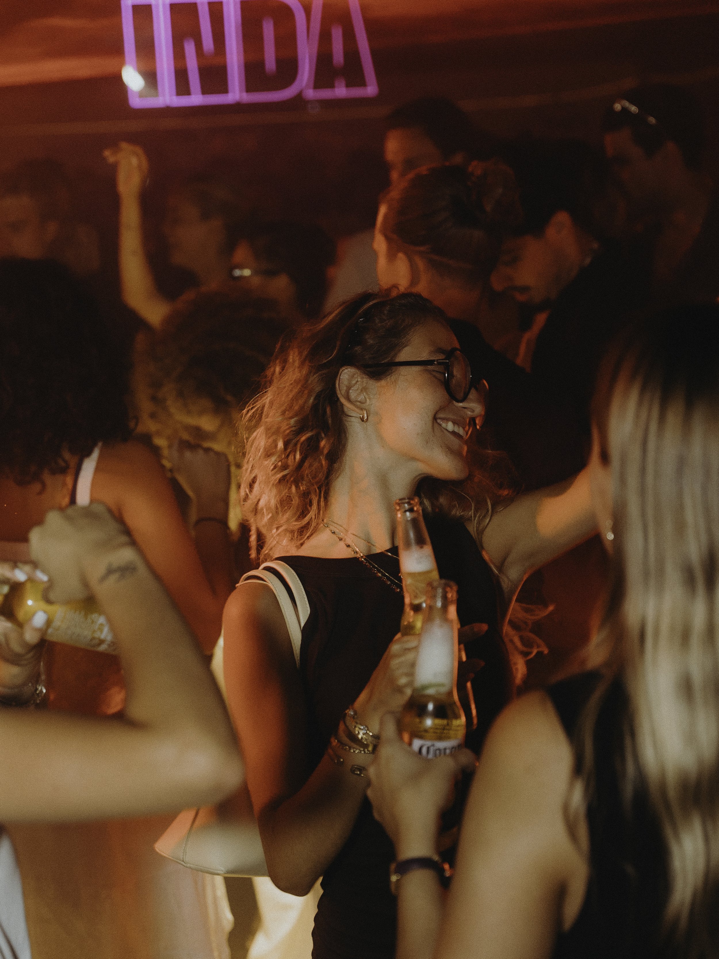 Fête dans une discothèque avec des personnes souriantes qui tiennent des bouteilles de bière Corona, ambiance lumineuse rouge et néons, des gens dansant.