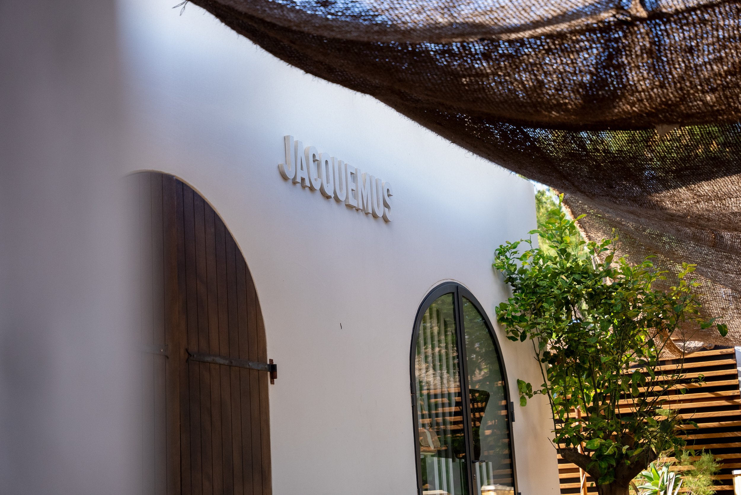 Façade d'un bâtiment blanc avec le nom 'JACQUEMUS' en lettres blanches, porte en bois arqué, une fenêtre en verre avec une grille noire, et un arbre vert à côté. Sombrage d'une toile de contrôle soleil en haut.