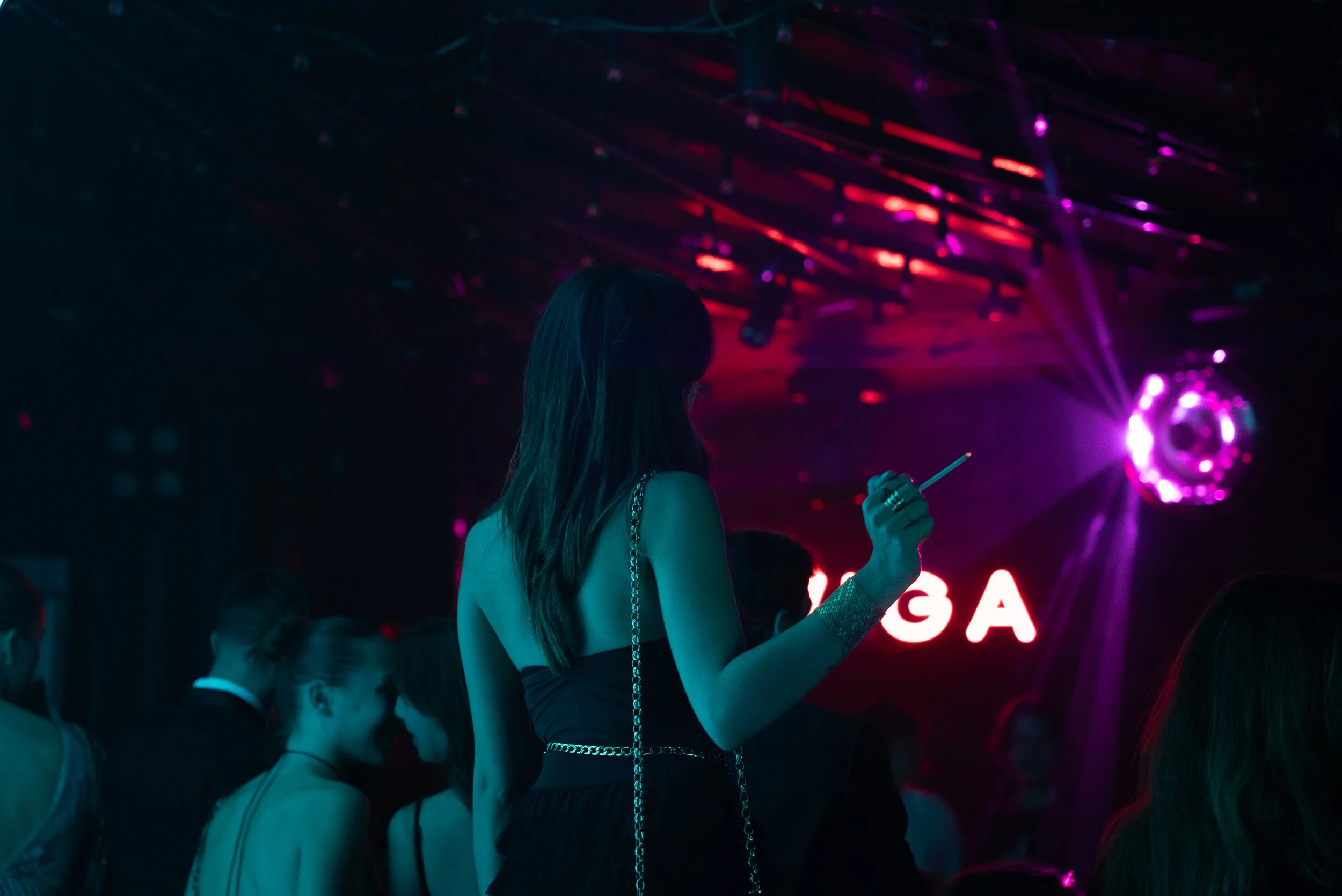 Une femme avec une robe noire et un sac à chaîne métallique, tenant une cigarette, dans une ambiance sombre avec des lumières violettes et rouges, au fond un panneau lumineux avec le mot 'GAGA'.
