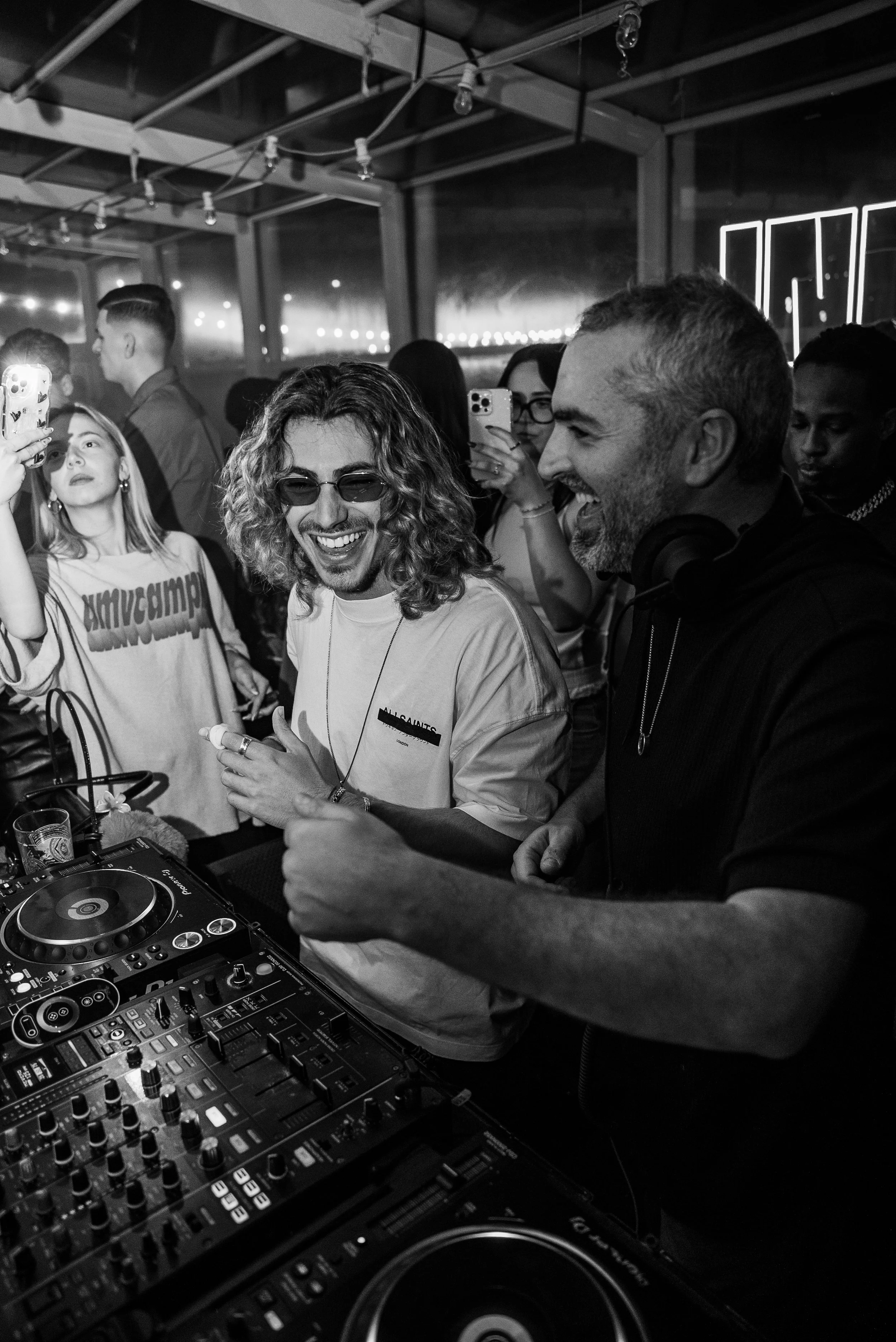 Deux hommes à une cabine de DJ, entourés de personnes dansant et prenant des photos lors d'une fête. L'ambiance est festive et lumineuse, avec des éclairages au fond.