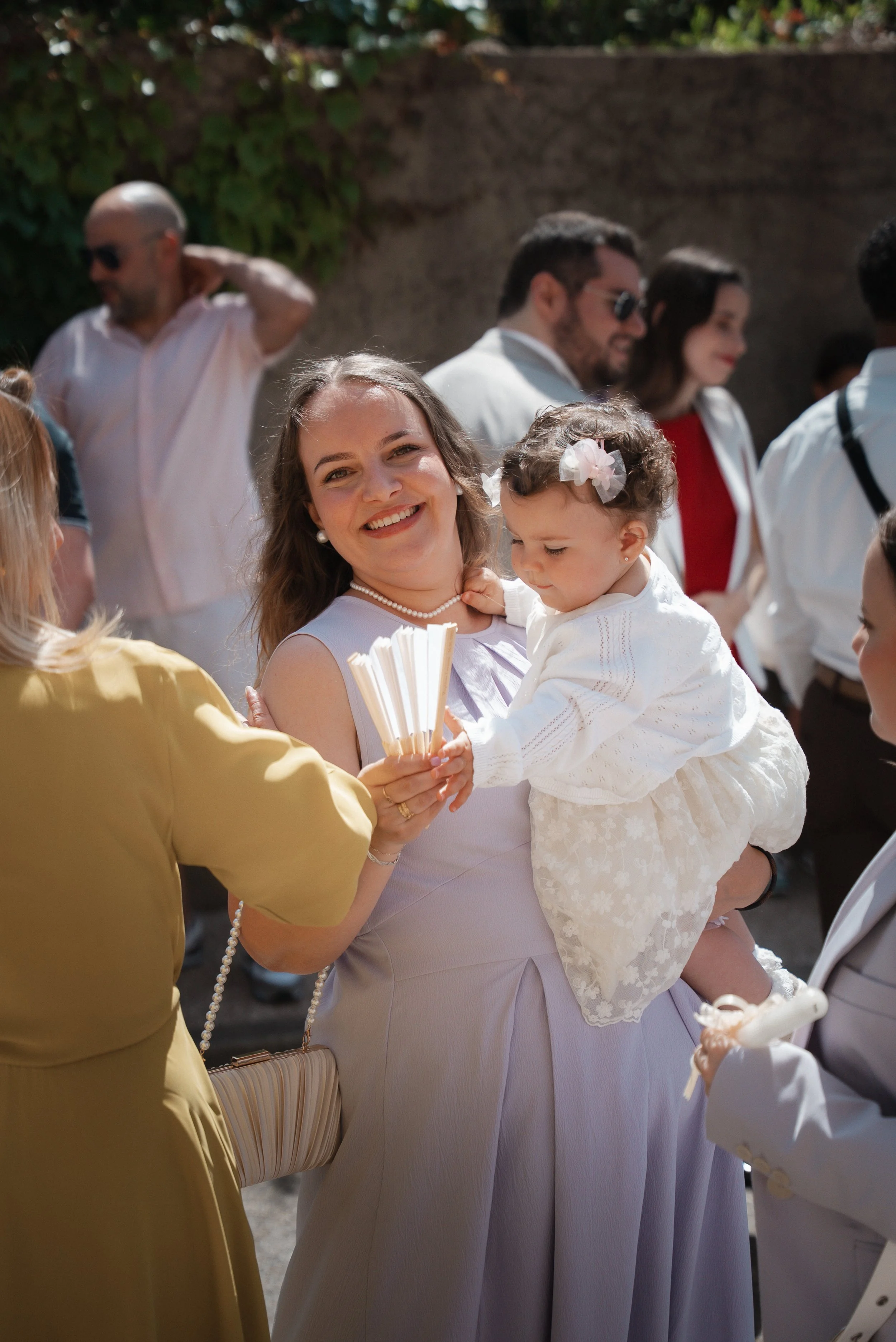 Groupe de personnes lors d'une célébration en plein air, avec une femme souriante tenant une enfant en robe blanche et une autre personne en jaune à côté.