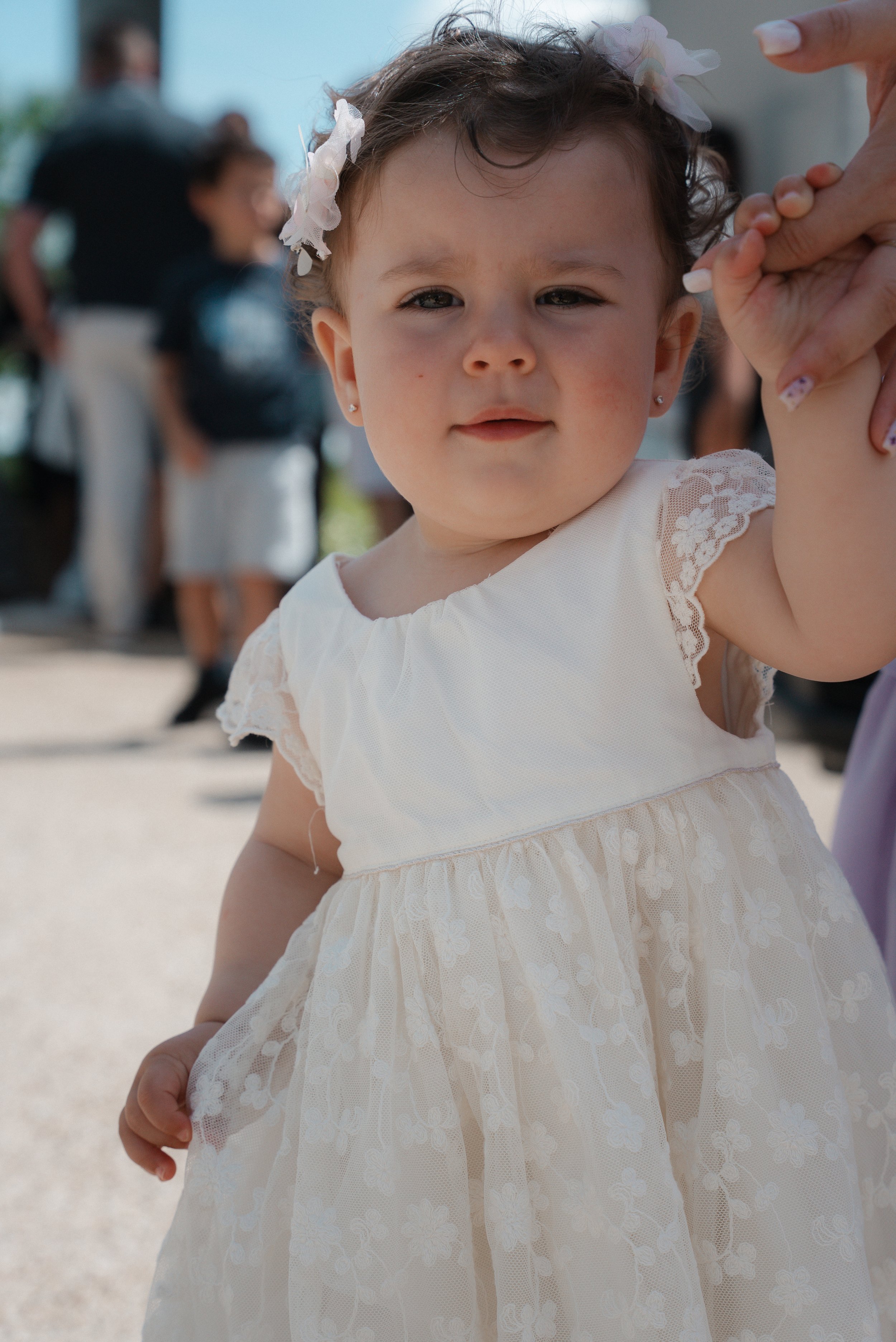 Jeune fille en robe blanche avec des fleurs dans les cheveux, tenant la main d'un adulte, lors d'un événement extérieur.