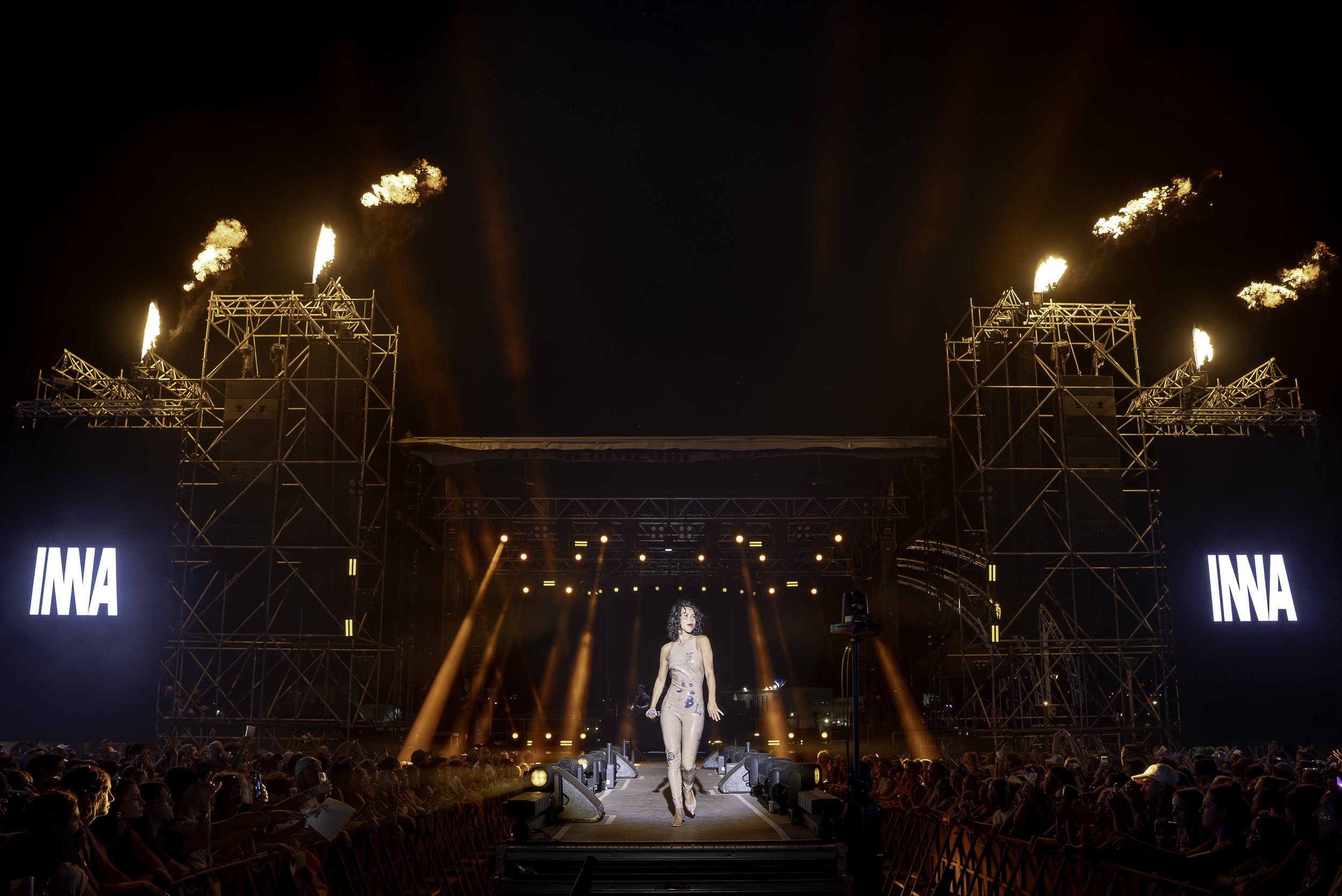 Une scène de concert ou de spectacle en plein air avec un grand public, un artiste féminine sur scène, un éclairage coloré et des flammes sur la structure du décor, avec deux grands écrans affichant le texte 'IMAA'.