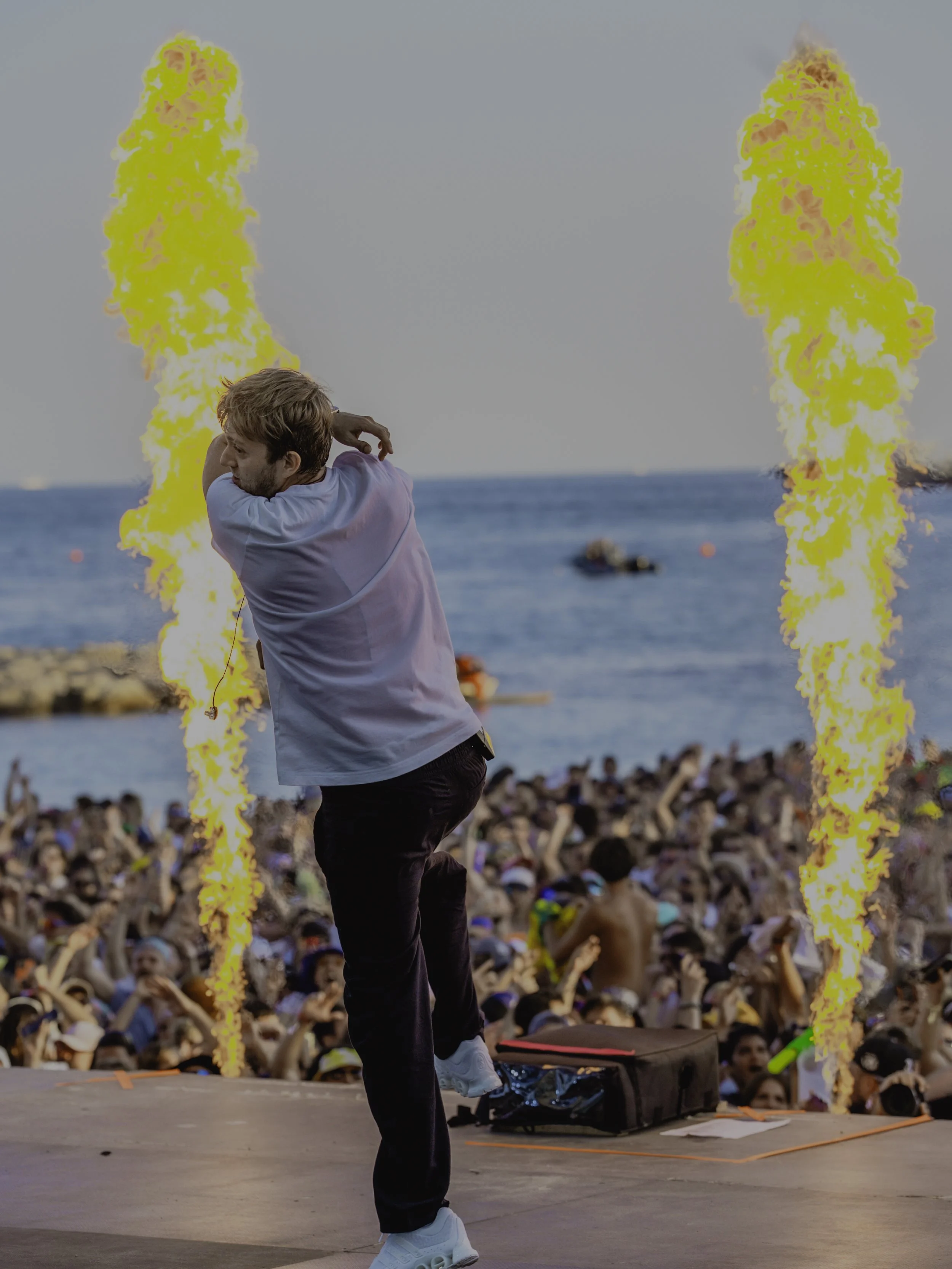 Un DJ sur scène lors d'un festival en bord de mer, avec deux flammes projetées en arrière-plan, un grand public en train de danser et de regarder l'artiste.