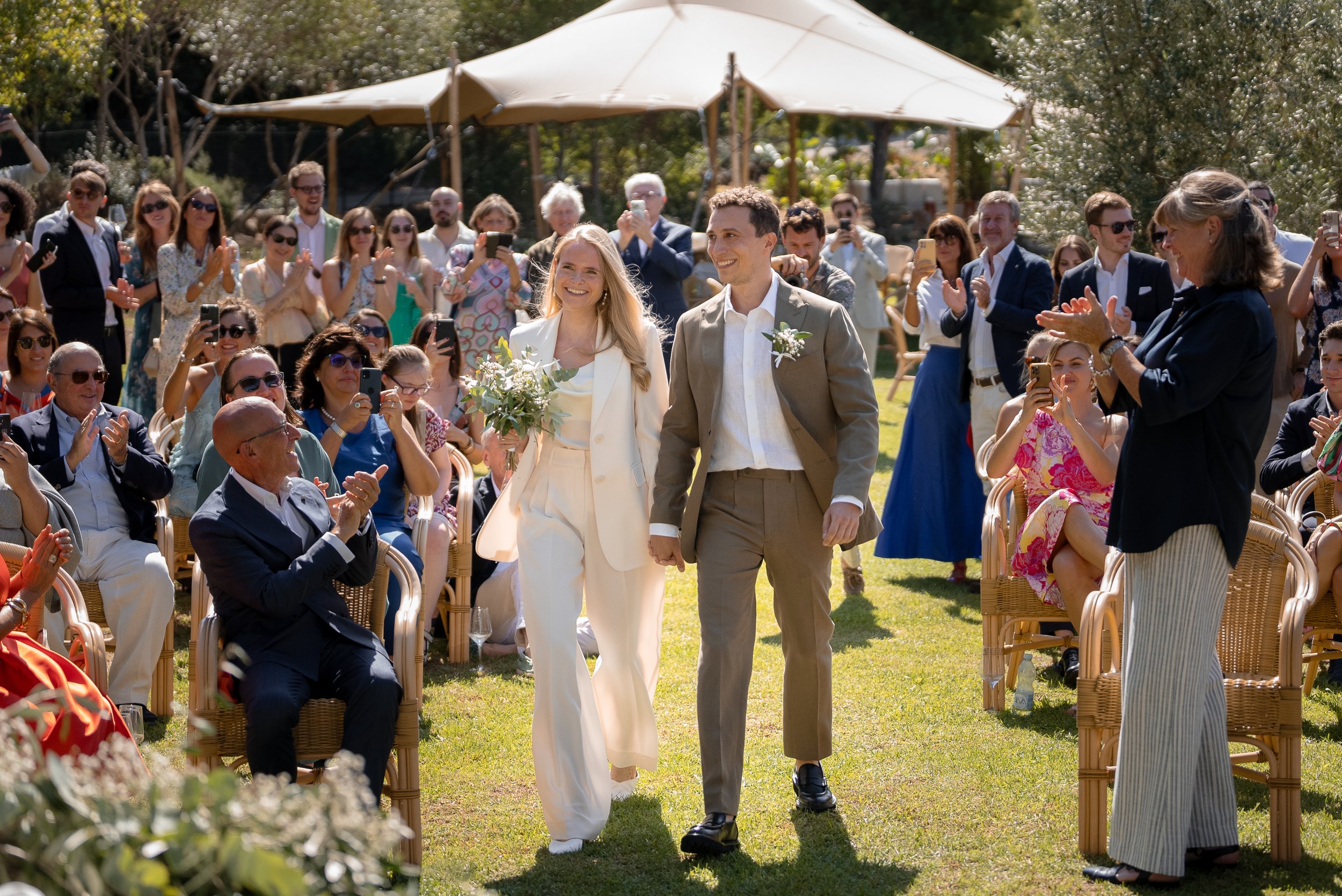 Un couple de mariés marche main dans la main lors d'une cérémonie en plein air, entouré d'invités applaudissant et prenant des photos, sous un grand parasol blanc.