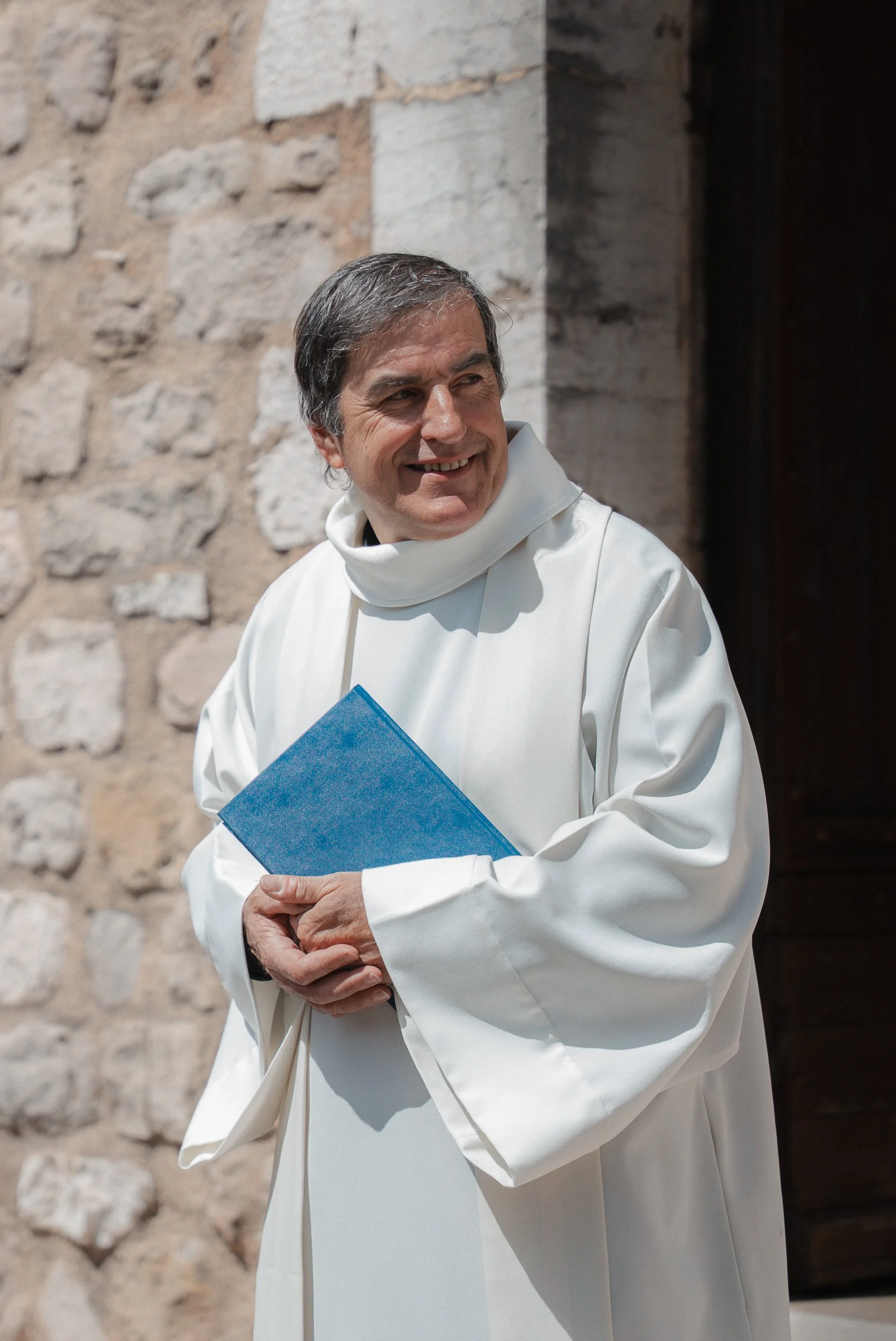 Un prêtre en robe blanche, tenant un livre bleu, souriant à l'extérieur devant un mur en pierre.